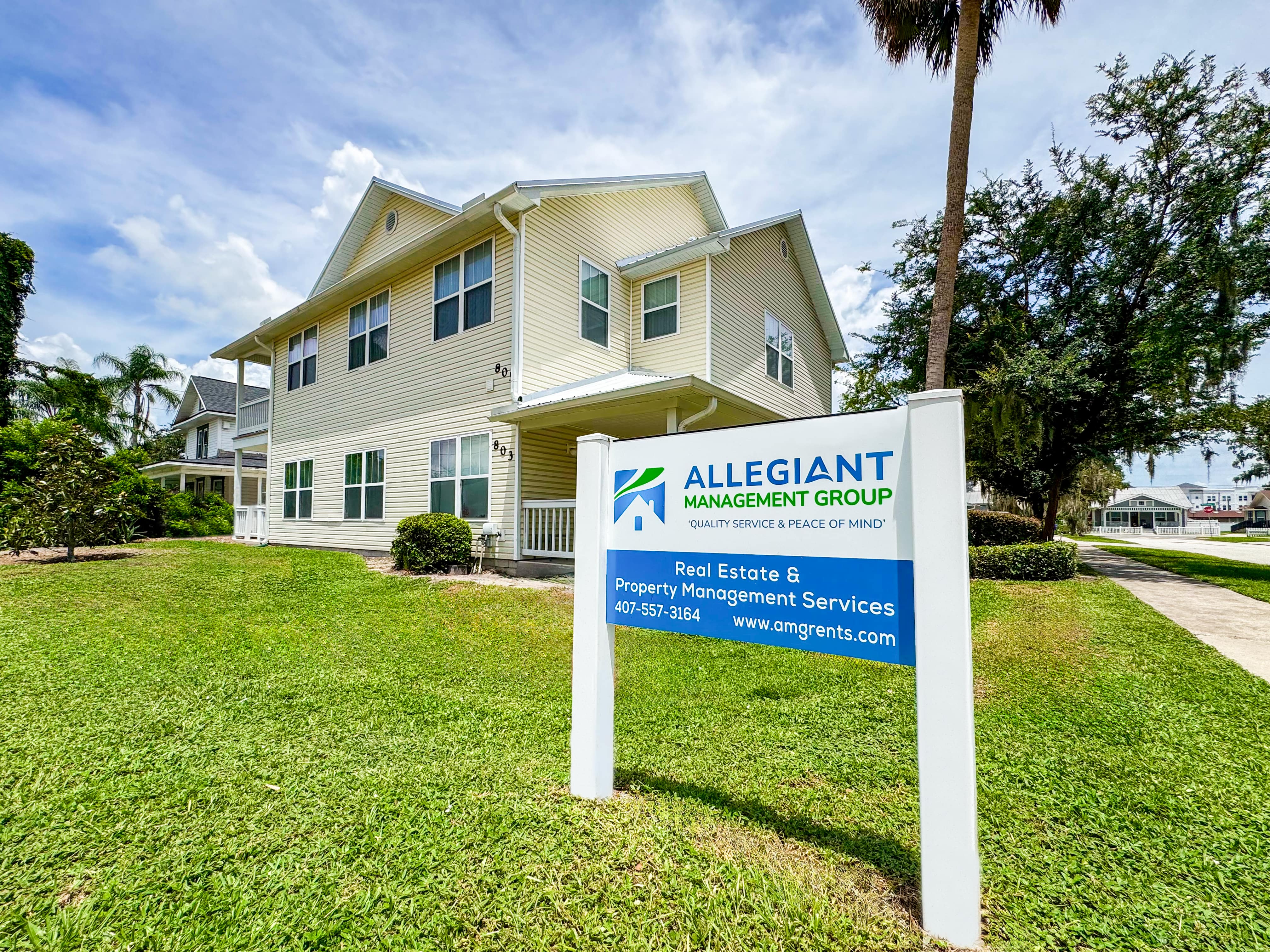 Exterior view of Allegiant Management Group’s office building, a two-story beige building with white trim, featuring a well-maintained lawn and a prominent blue and green company sign displaying their real estate and property management services.