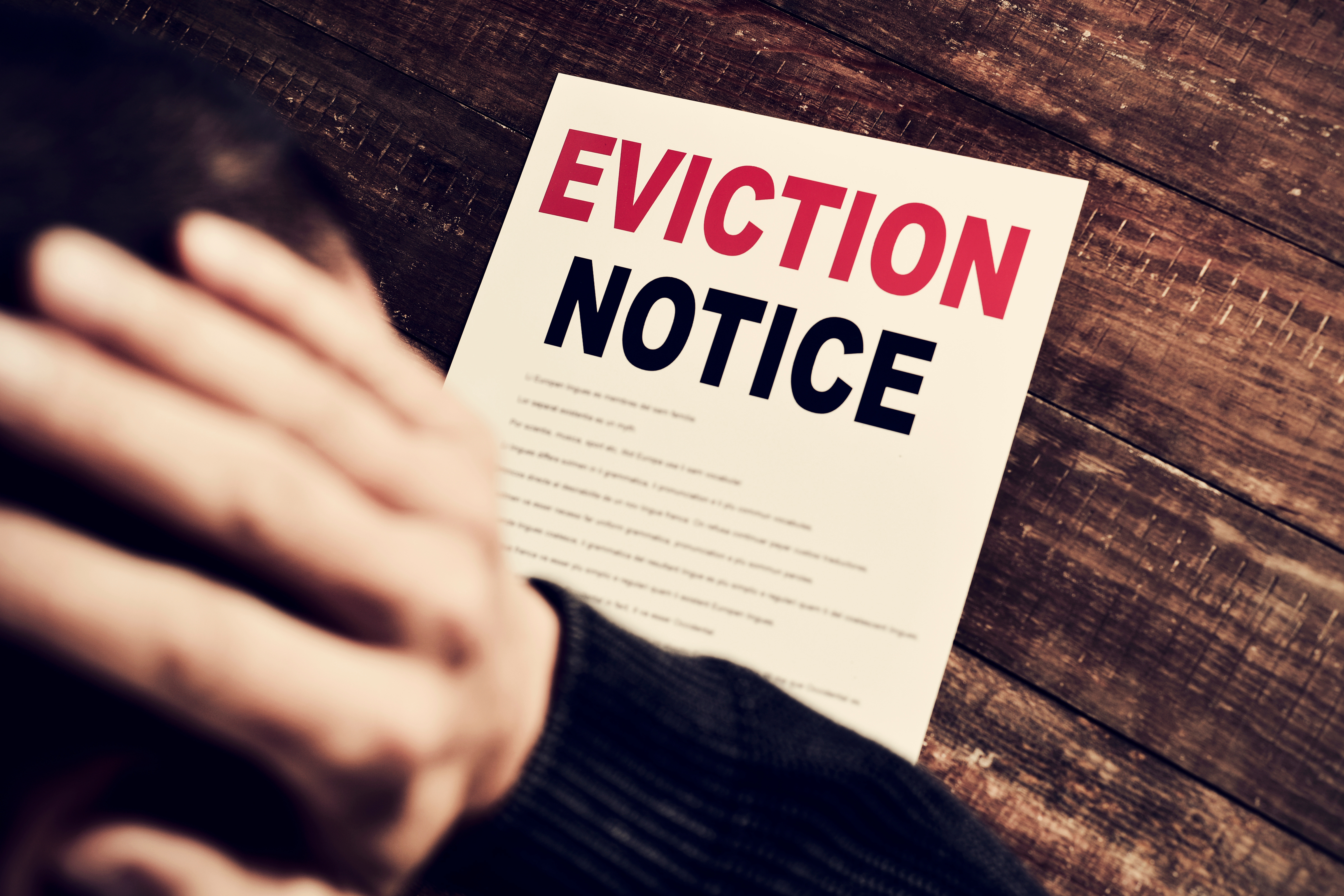 A person holding their head in distress with an eviction notice on a wooden table in the background.