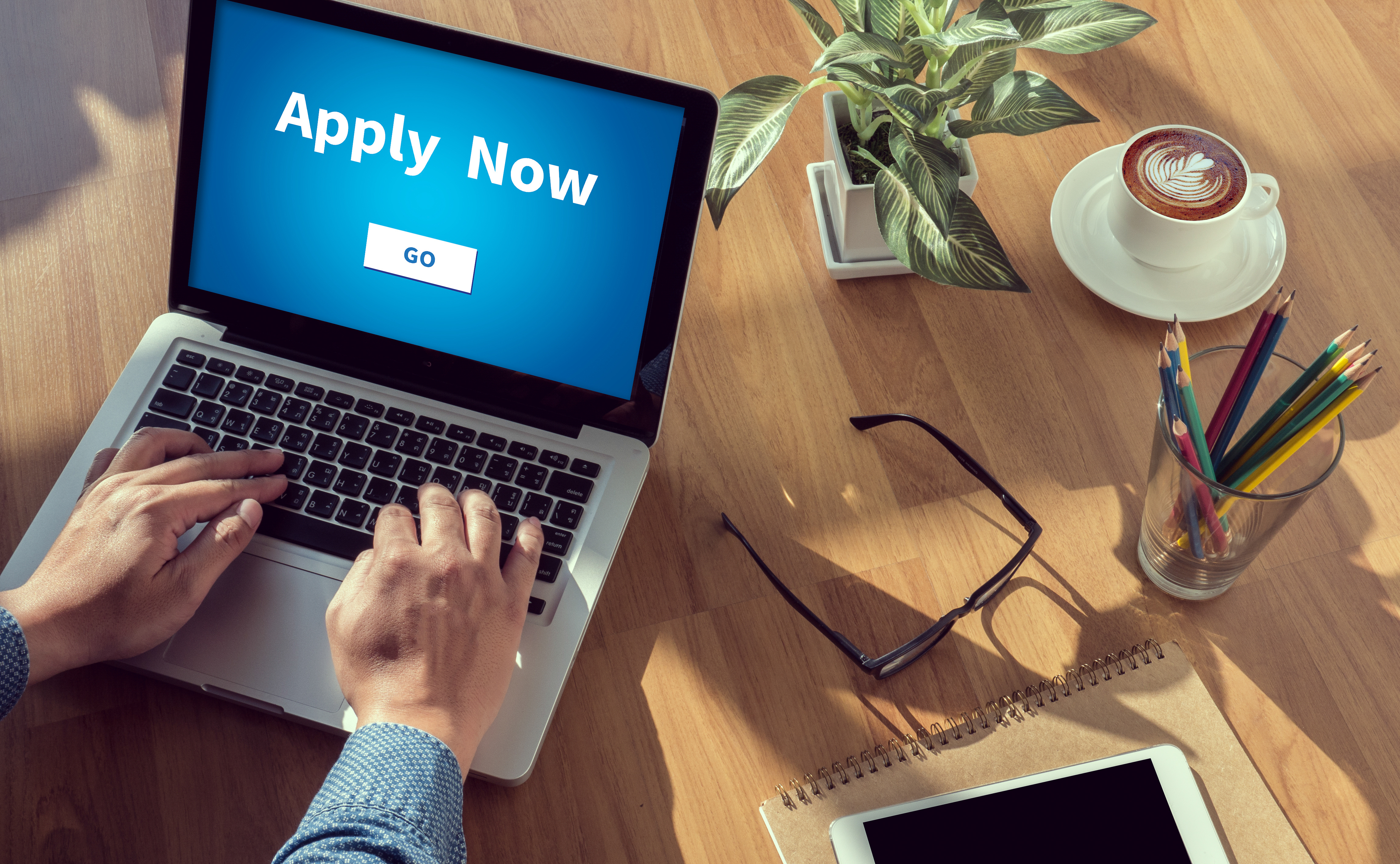 A laptop screen displaying “Apply Now” on a table surrounded by office supplies, a plant, and a cup of coffee.