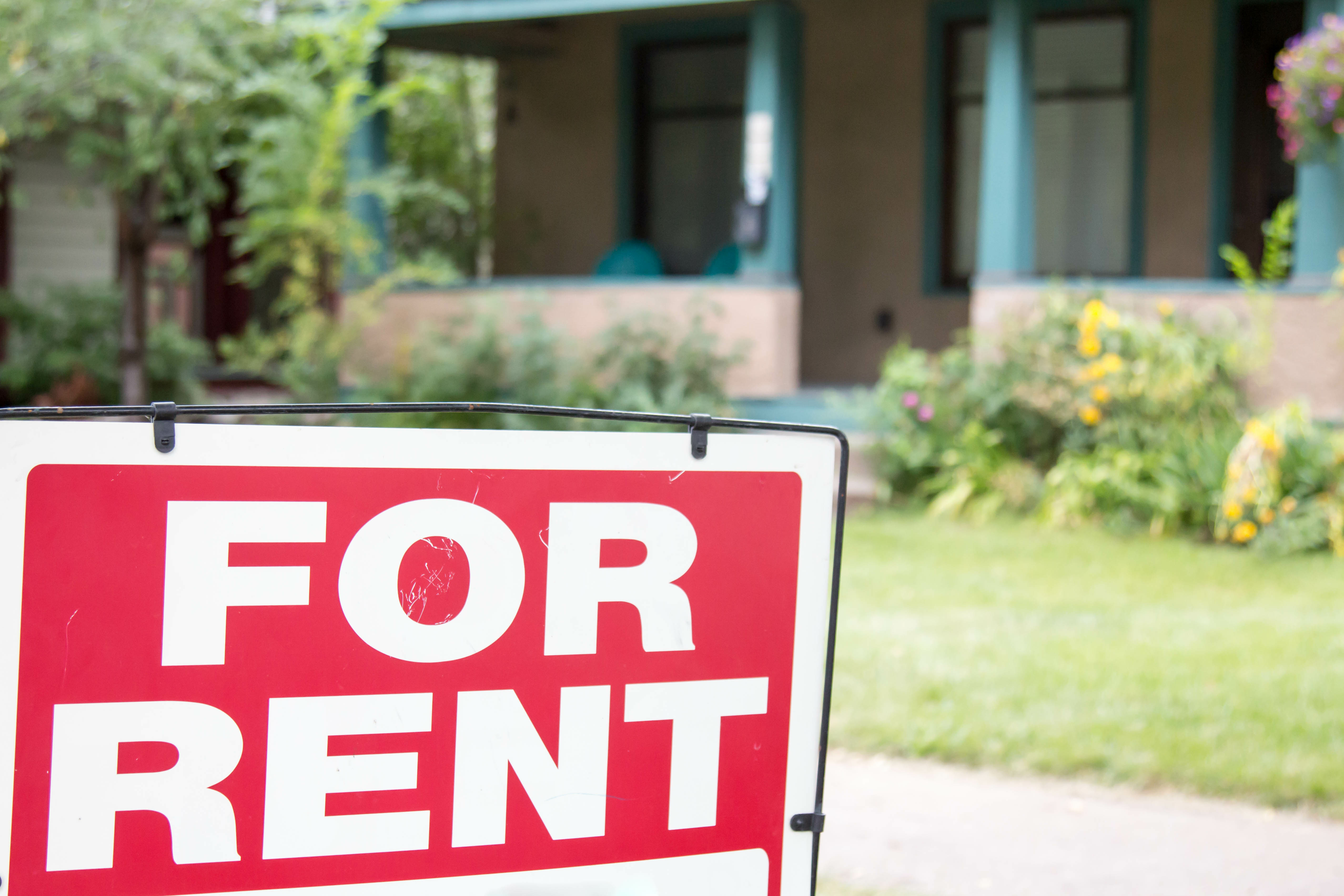 Close-up of a ‘For Rent’ sign in front of a house with greenery and flowers in the background.