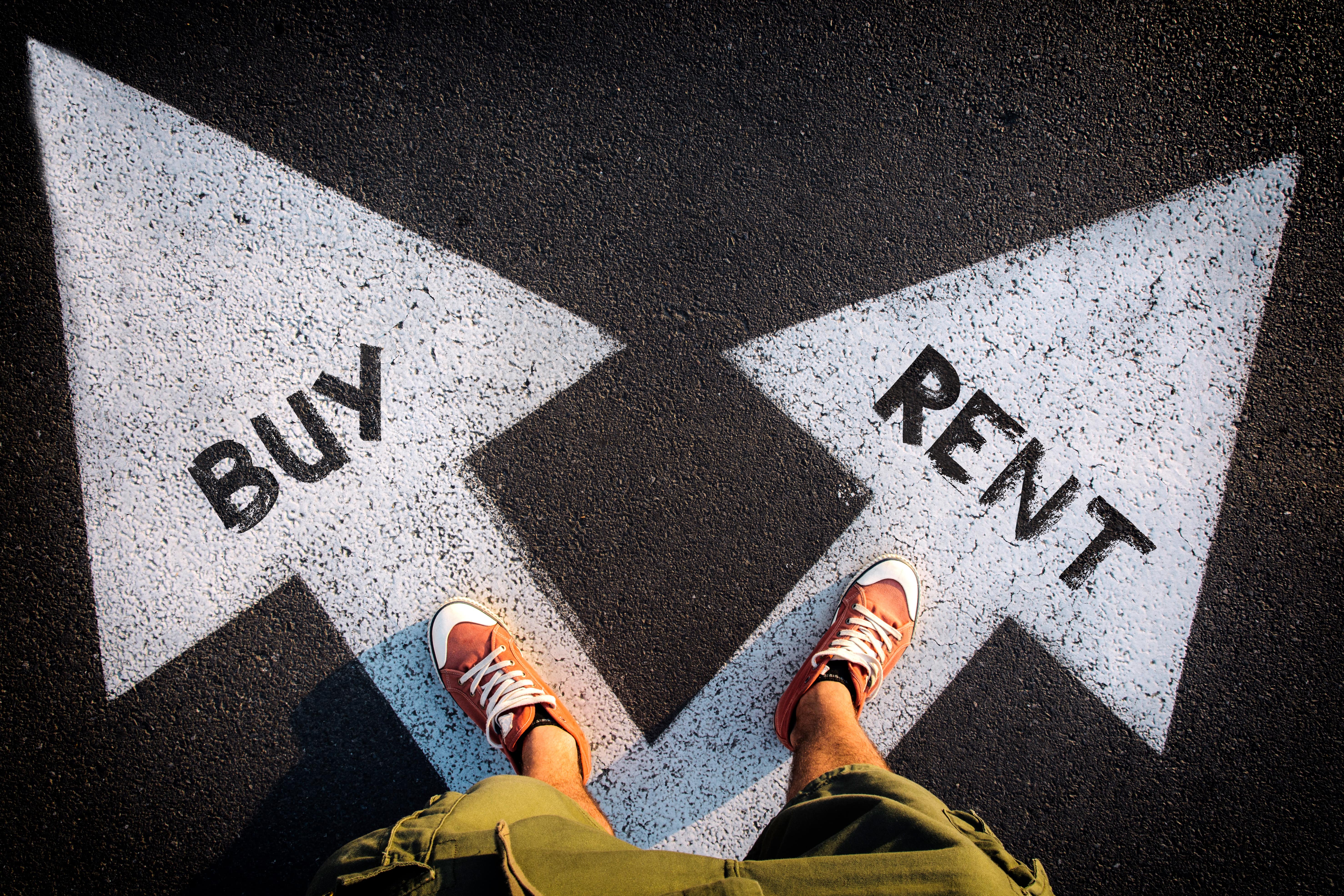 A person wearing orange sneakers stands at a fork in the road with two large white arrows painted on the pavement. One arrow points to 'BUY,' and the other points to 'RENT,' symbolizing a decision between purchasing or renting a home.