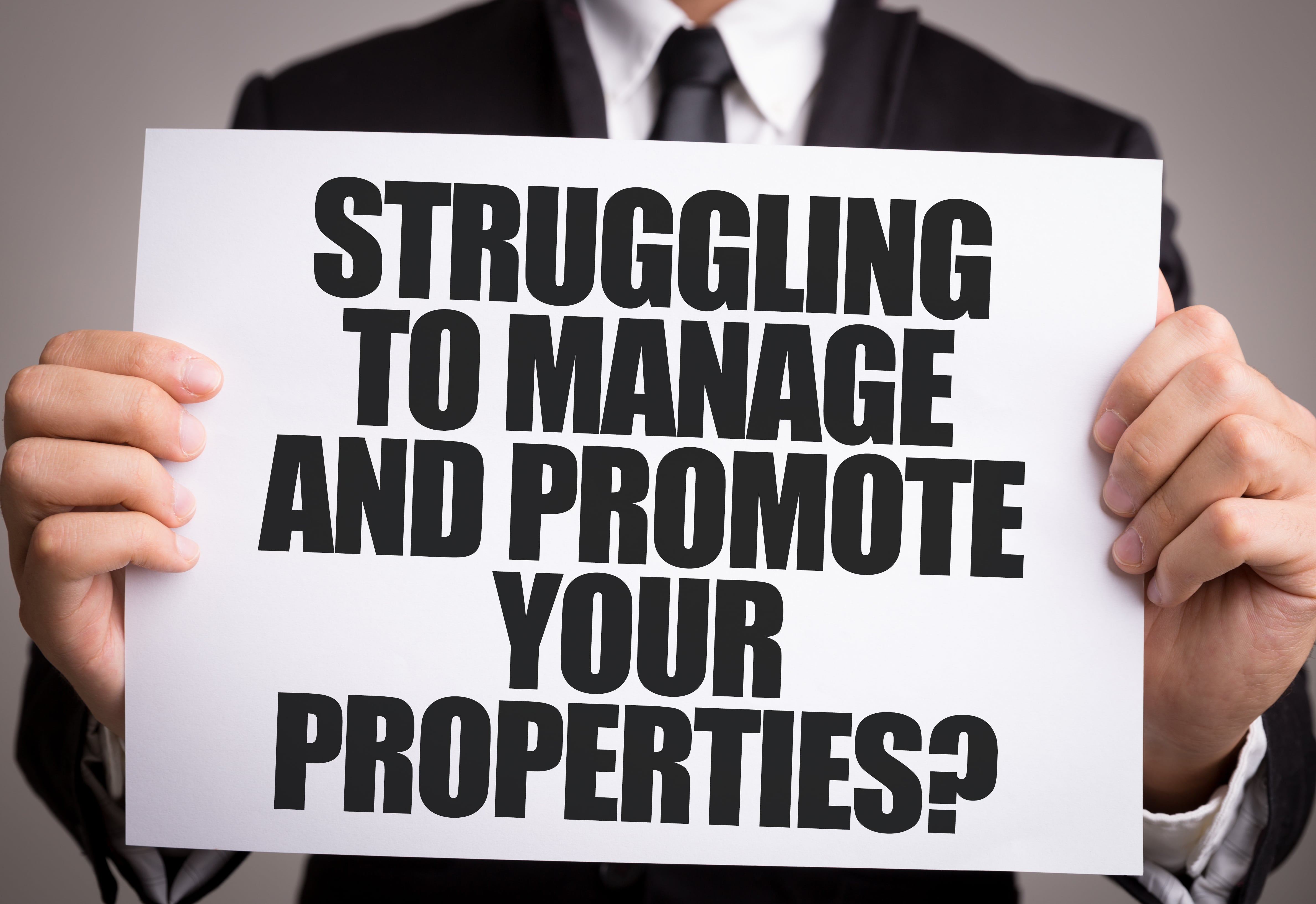 A person in a business suit holding a white sign with bold black text that reads, 'STRUGGLING TO MANAGE AND PROMOTE YOUR PROPERTIES?' The message highlights property management challenges and the need for professional assistance.