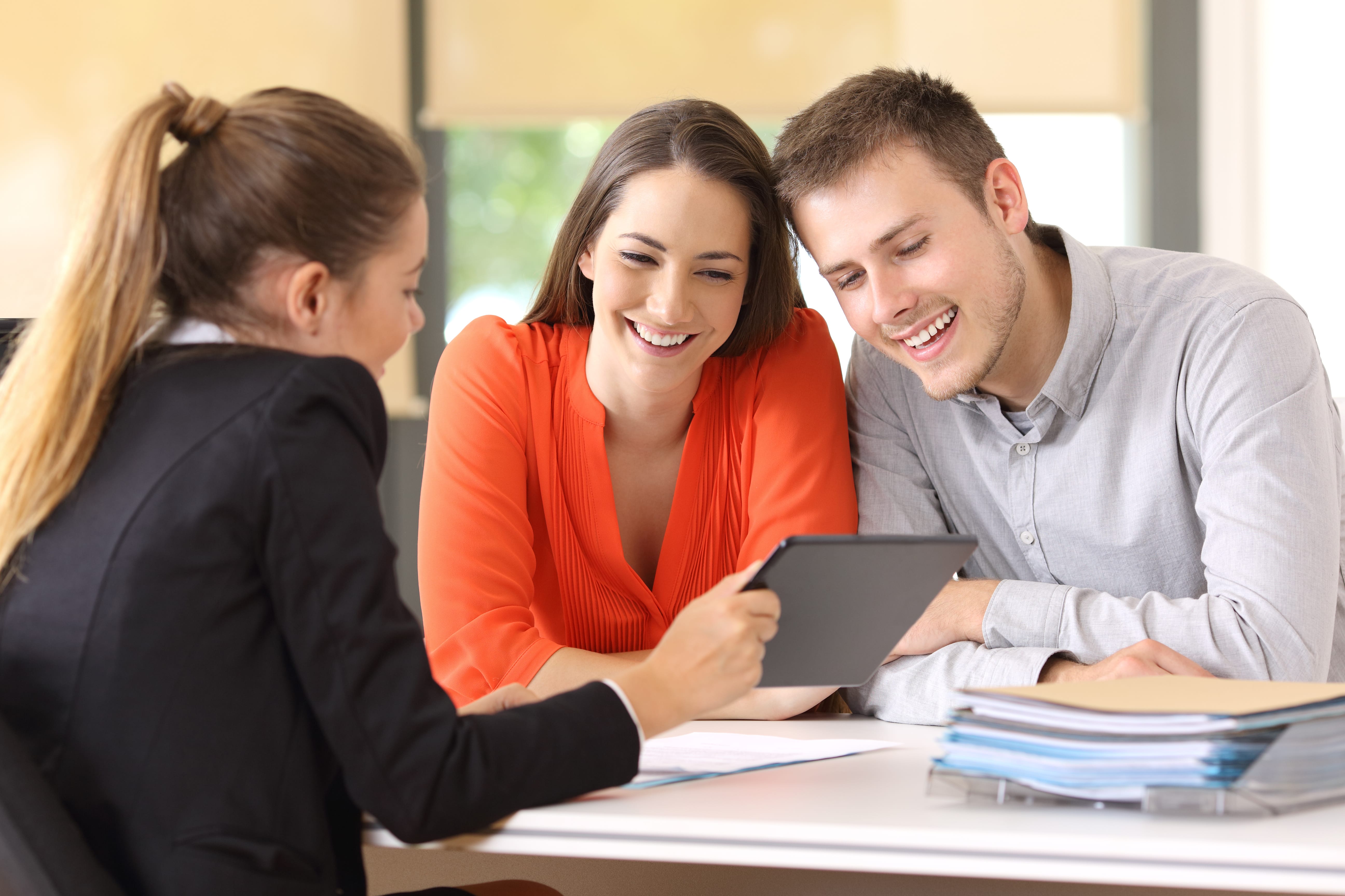 A real estate agent presenting documents on a tablet to a smiling couple in an office setting.