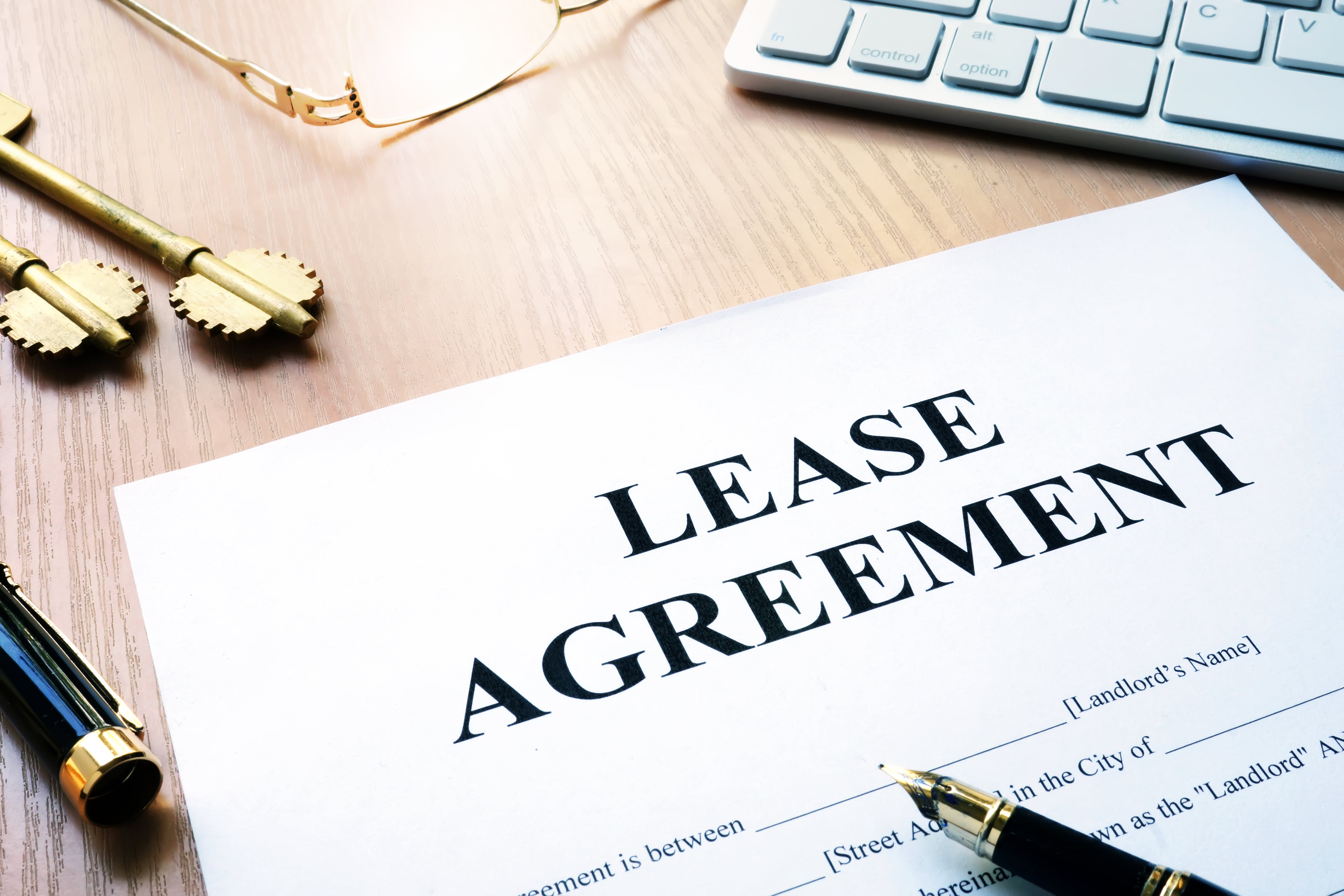 Lease agreement document on a wooden desk with gold keys, a fountain pen, and a computer keyboard, representing rental contracts and landlord-tenant agreements.