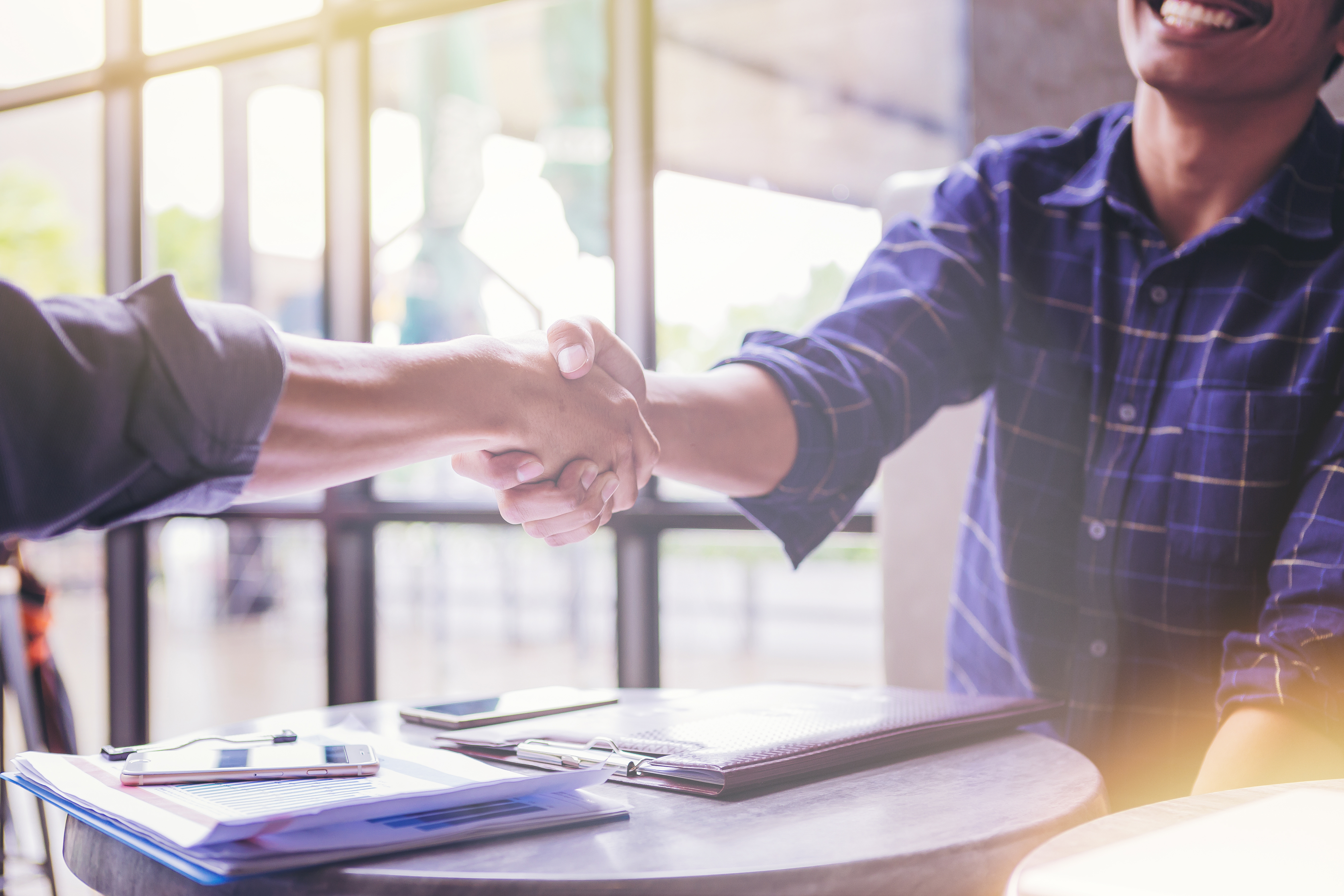 Two individuals shaking hands across a table, symbolizing an agreement or partnership. Documents, pens, and a smartphone are visible on the table.