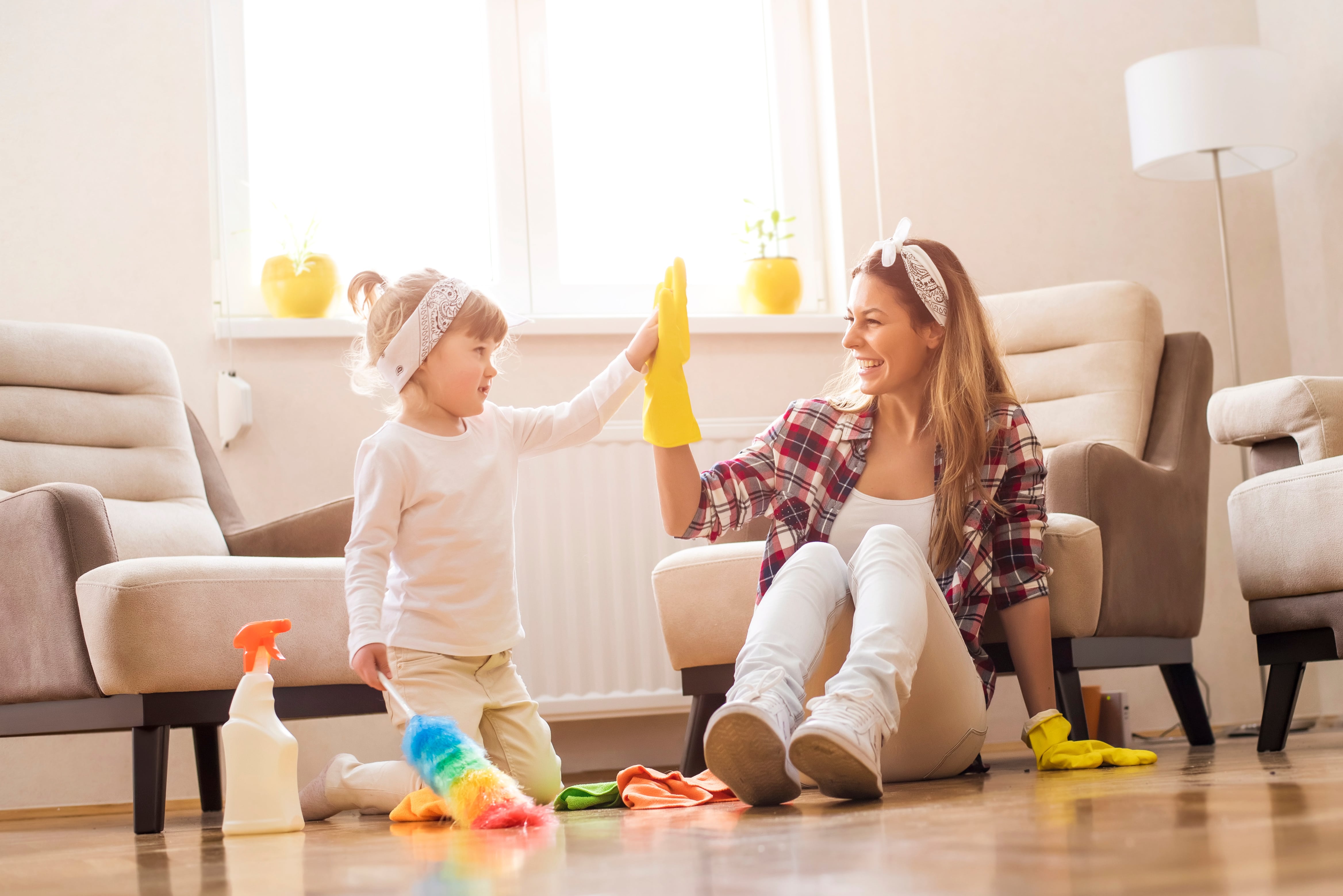 Mother and daughter high-five while spring cleaning their home, surrounded by cleaning supplies and a bright, tidy living room.