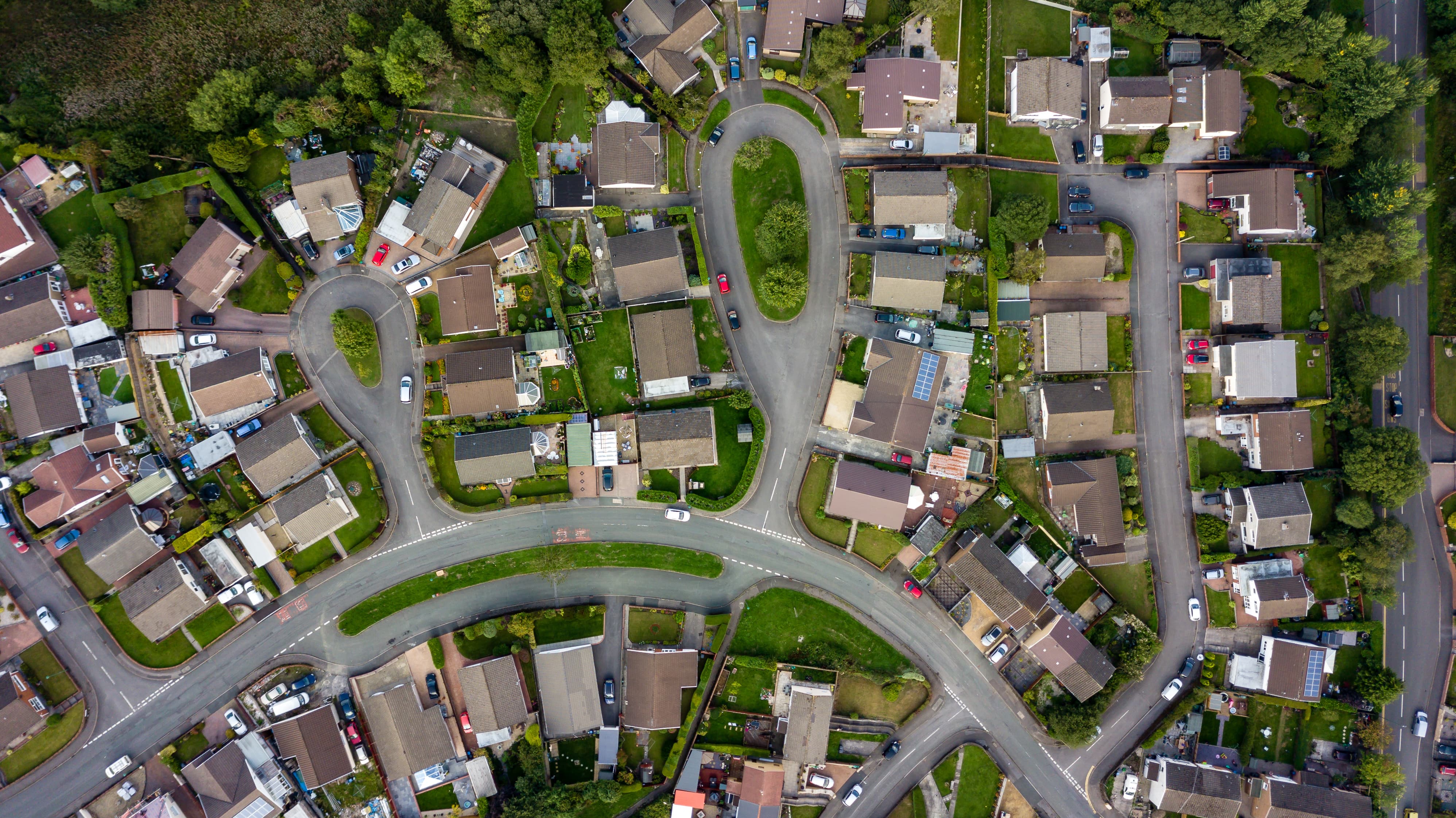 Aerial view of a suburban neighborhood with winding streets, single-family homes, green lawns, and parked cars, showing a well-planned residential community.