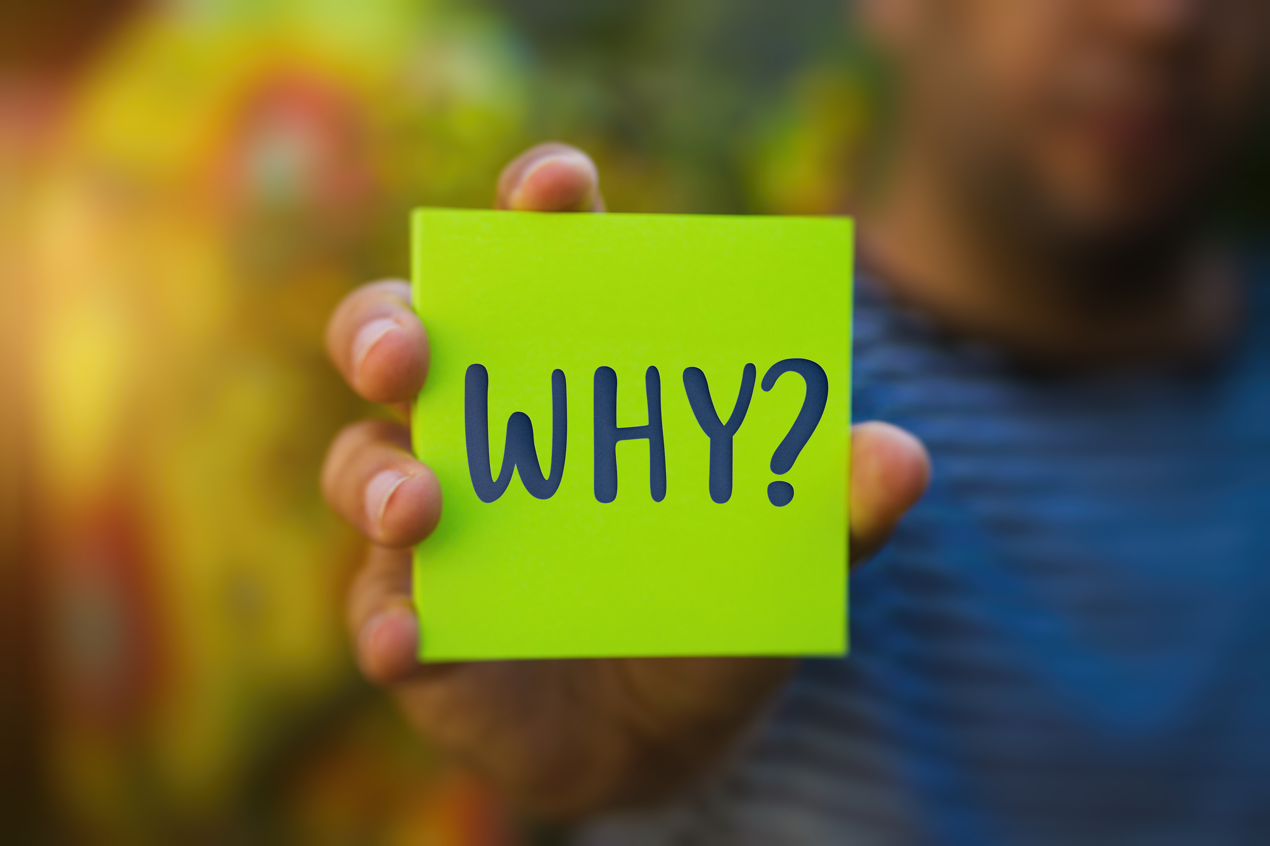 Close-up of a hand holding a green note with the word ‘WHY?’ written on it, symbolizing the importance of questioning and understanding decisions.