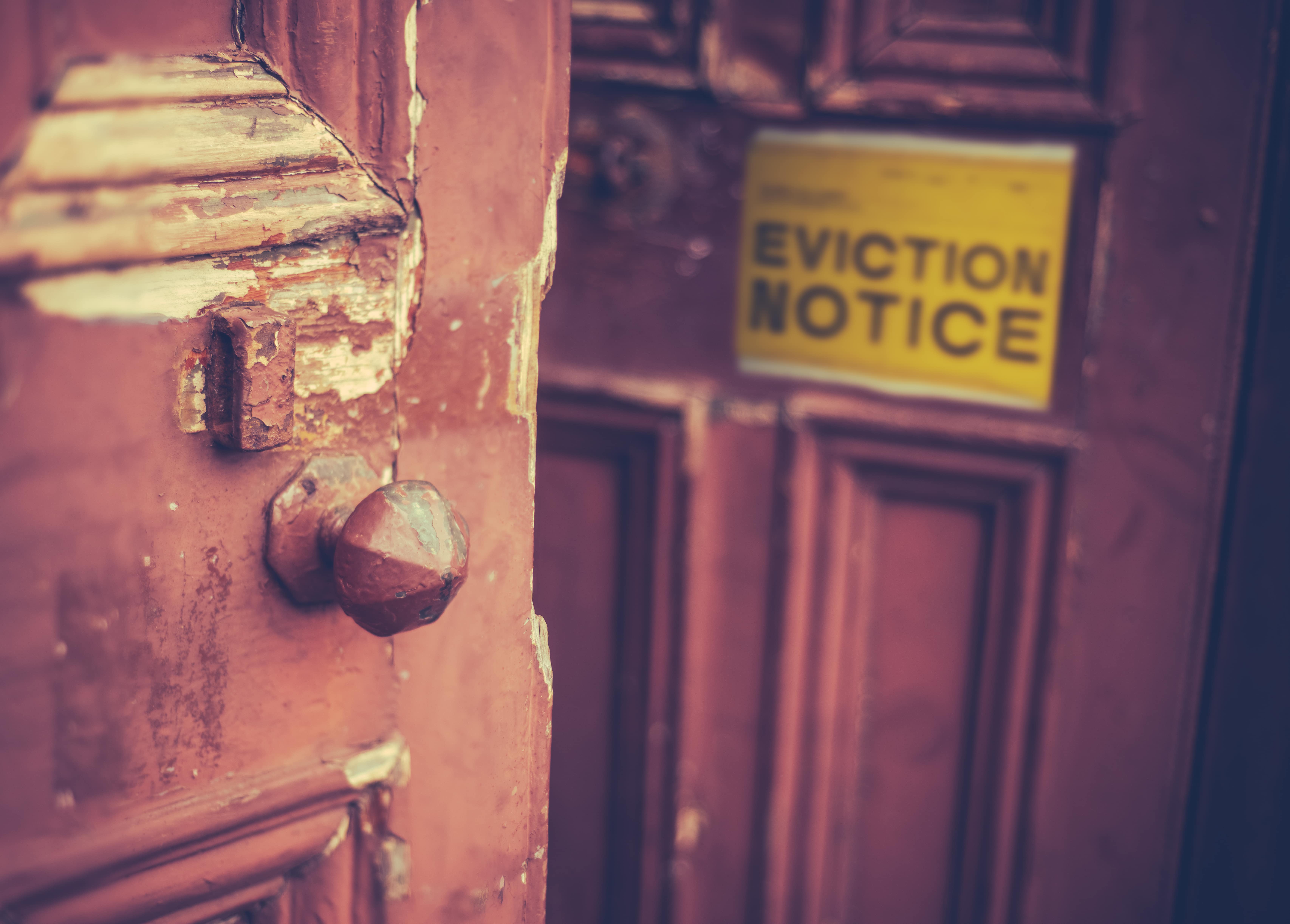 A close-up of a weathered wooden door with peeling paint, a rusty doorknob, and a blurred yellow eviction notice in the background.