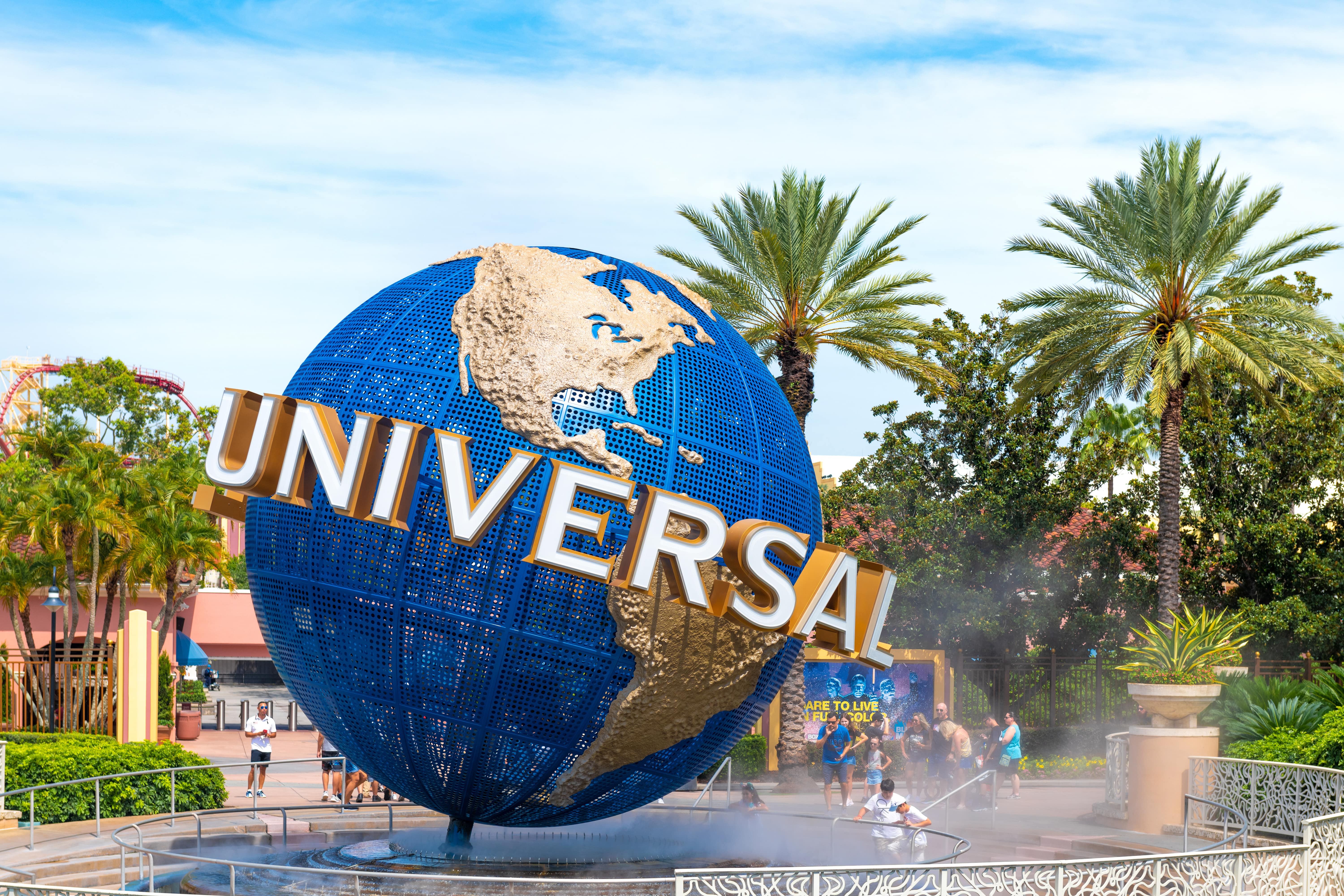 The famous blue and gold spinning globe with 'Universal' in bold letters, positioned at the entrance of Universal Orlando Resort. Palm trees and mist from a nearby fountain surround the globe, with visitors enjoying the theme park in the background.