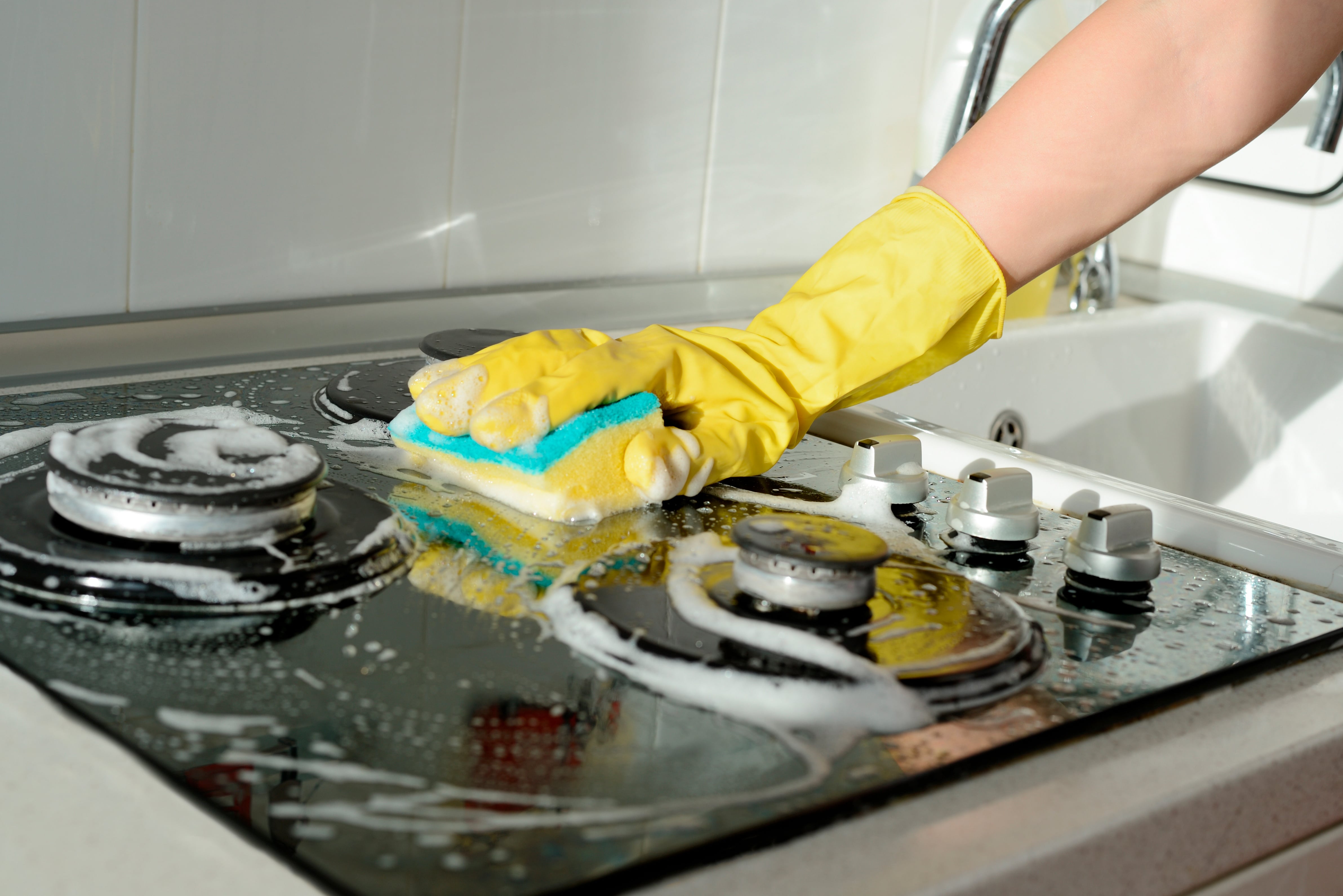 A person wearing yellow rubber gloves uses a sponge to scrub a stovetop covered in soap suds. The image represents cleaning tasks, likely related to property maintenance or move-in/move-out processes.