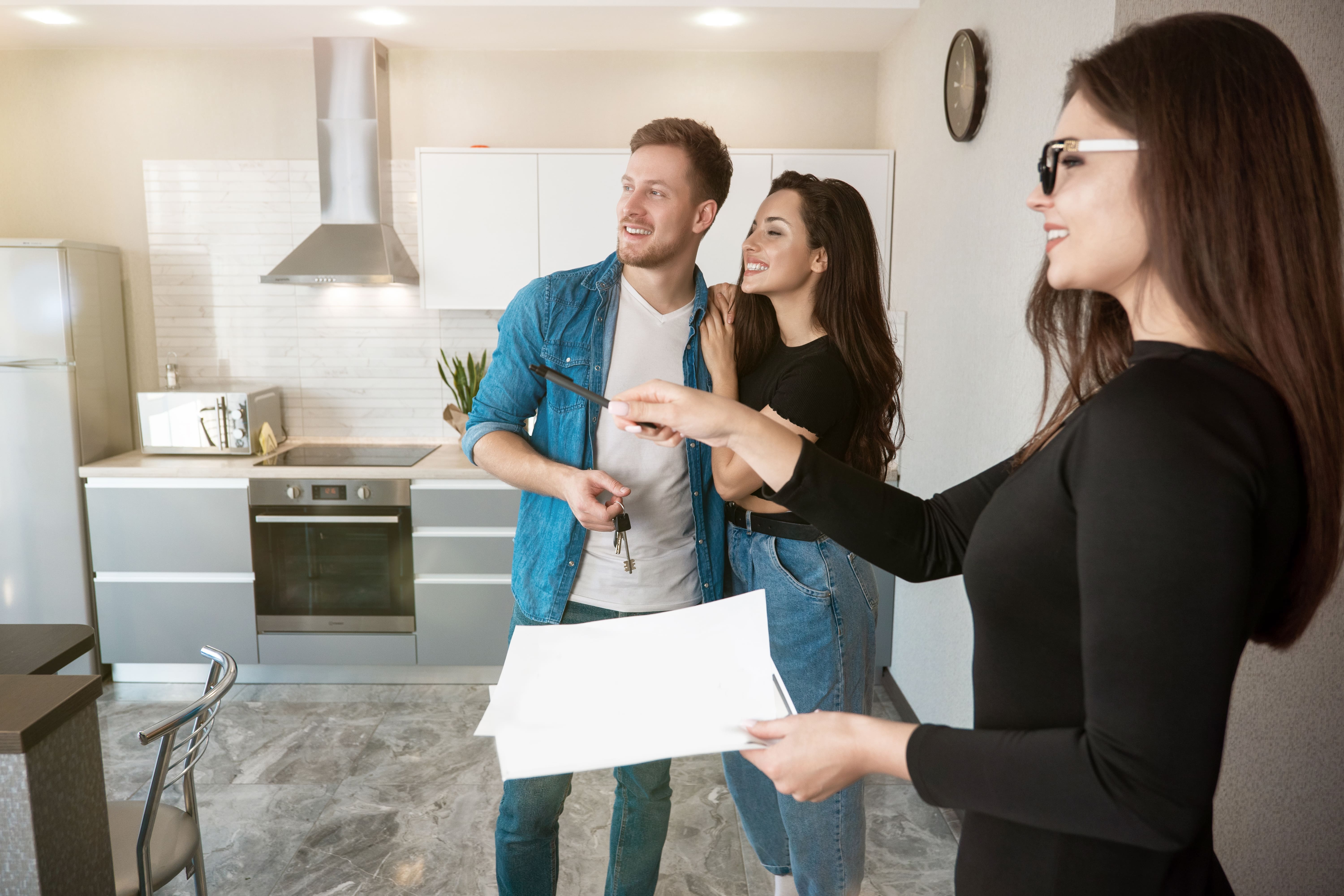 A real estate agent in a black outfit shows a modern kitchen to a happy couple holding new house keys.