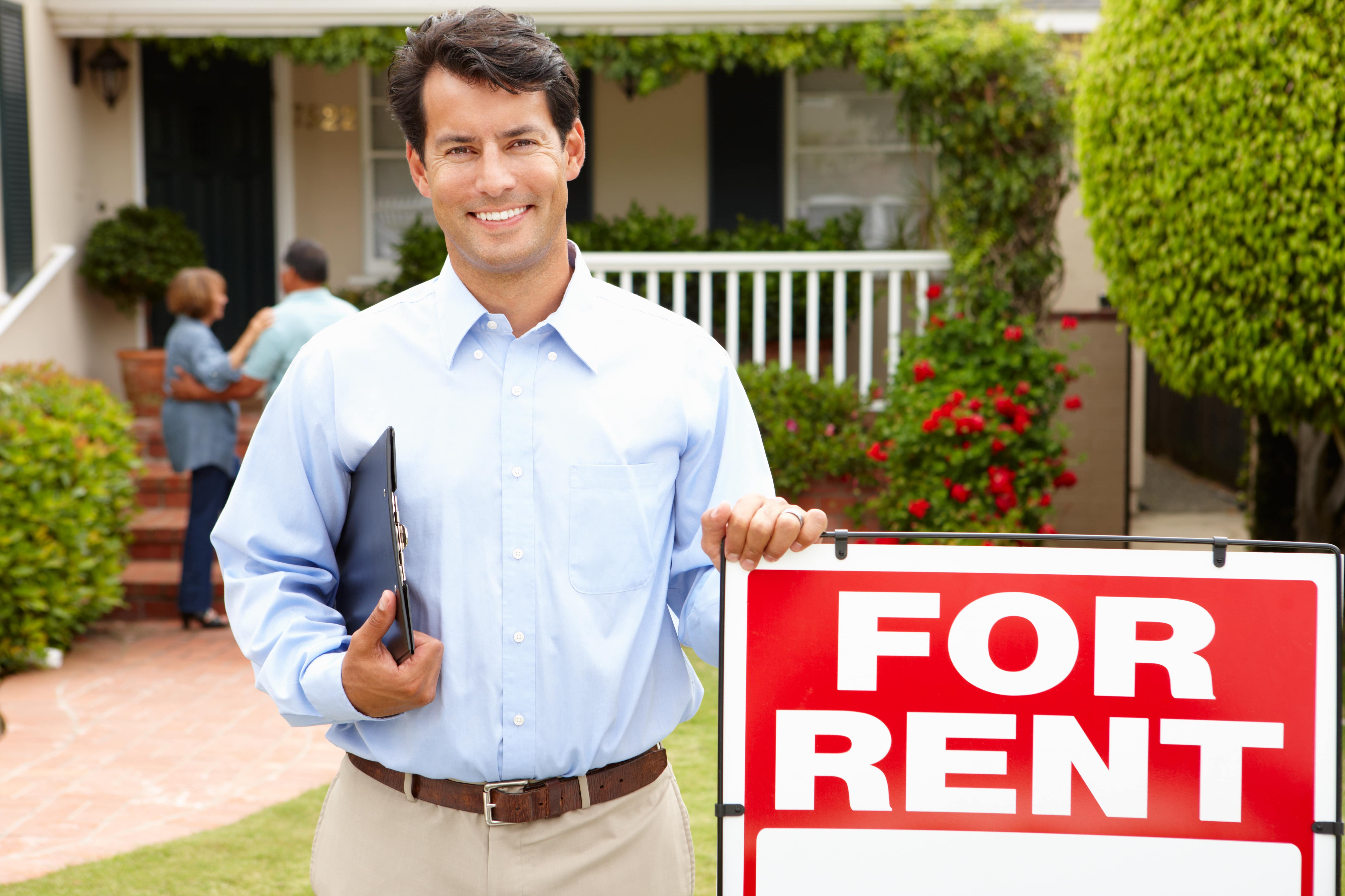 A smiling property manager standing next to a 'For Rent' sign in front of a rental property, representing property leasing.