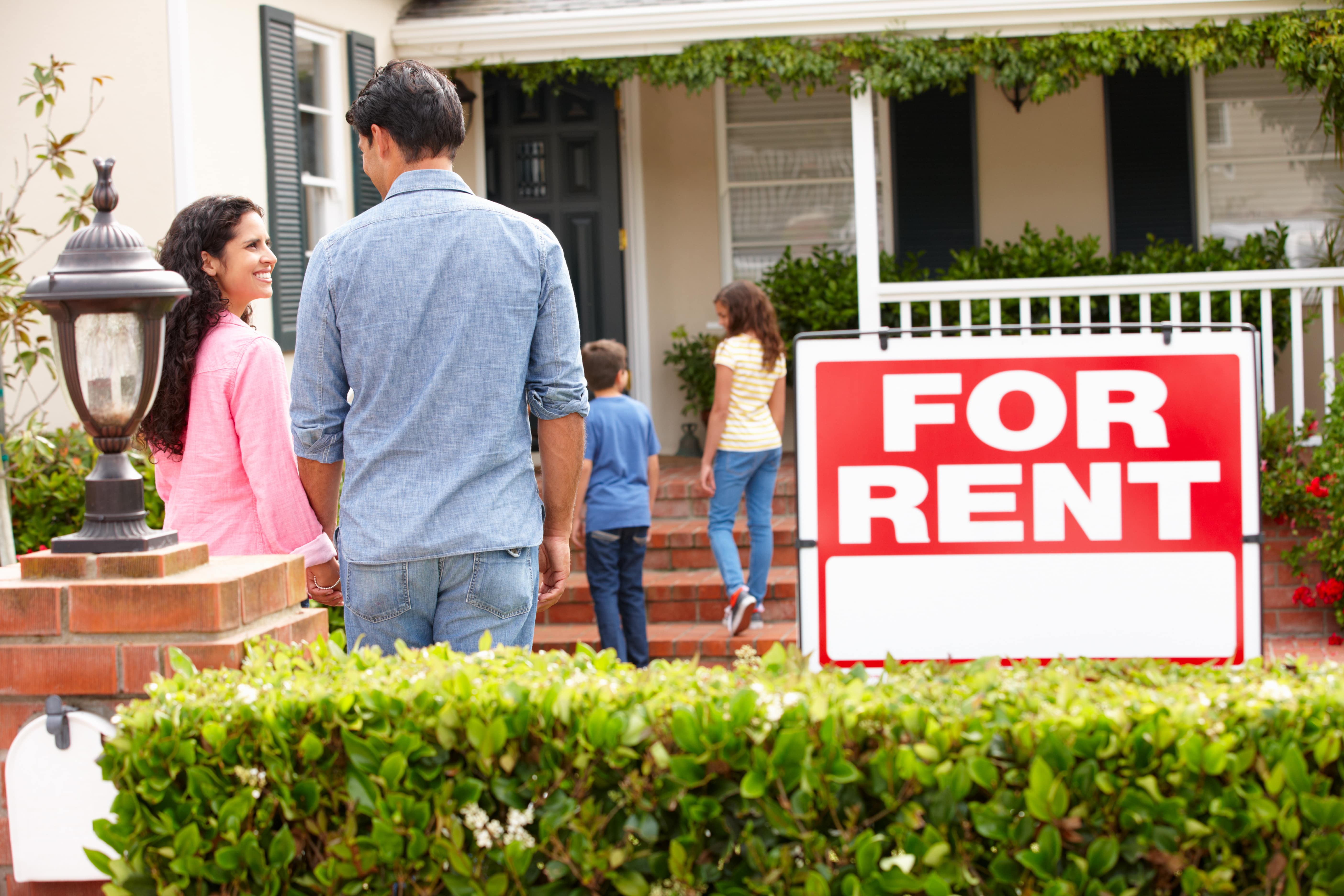 A family of four standing in front of a house with a 'For Rent' sign, discussing rental opportunities.