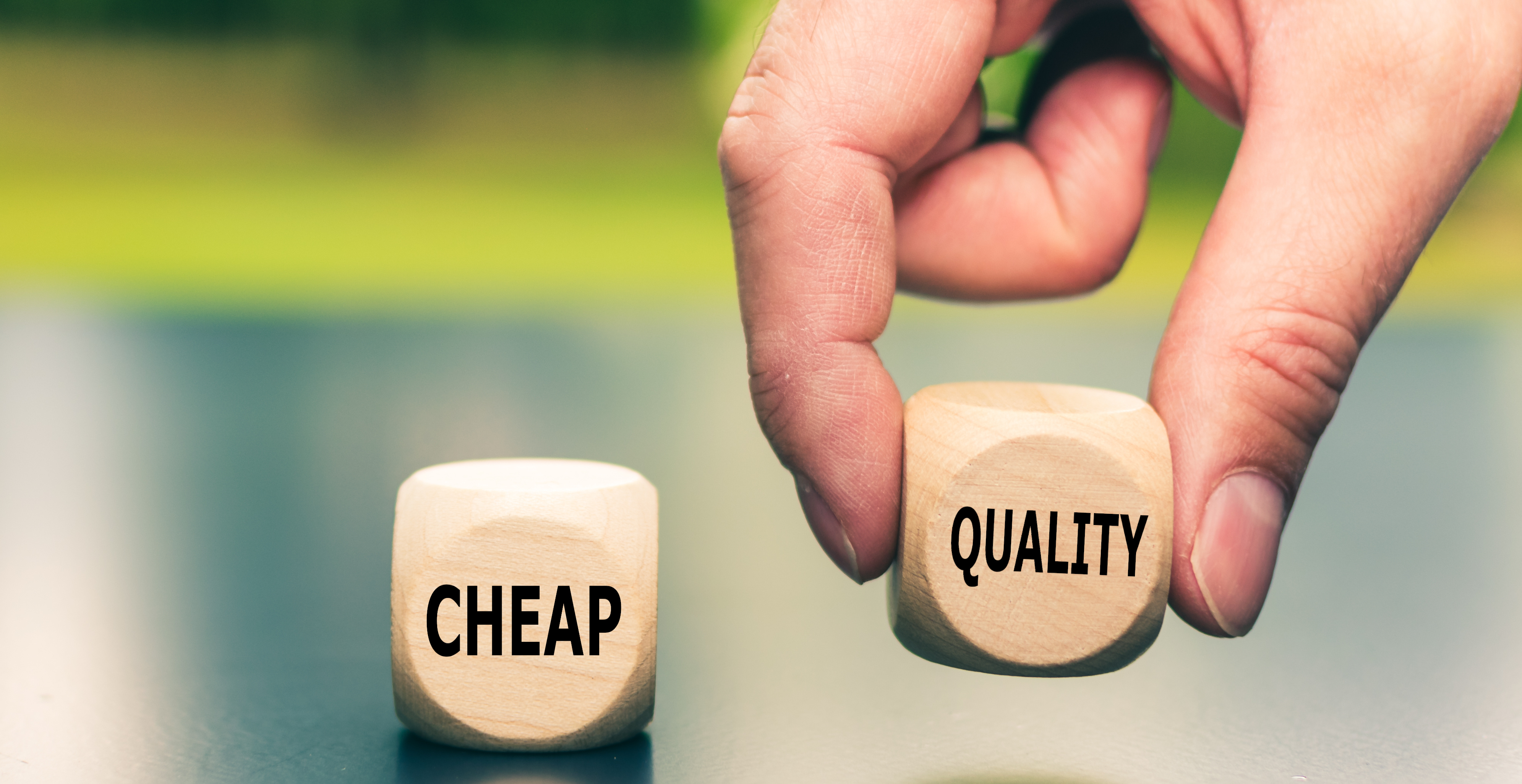 A hand is picking up a wooden block labeled “Quality,” while another block labeled “Cheap” remains on the table. The background is soft and blurred, indicating a decision between choosing cost-saving options versus quality products or services.