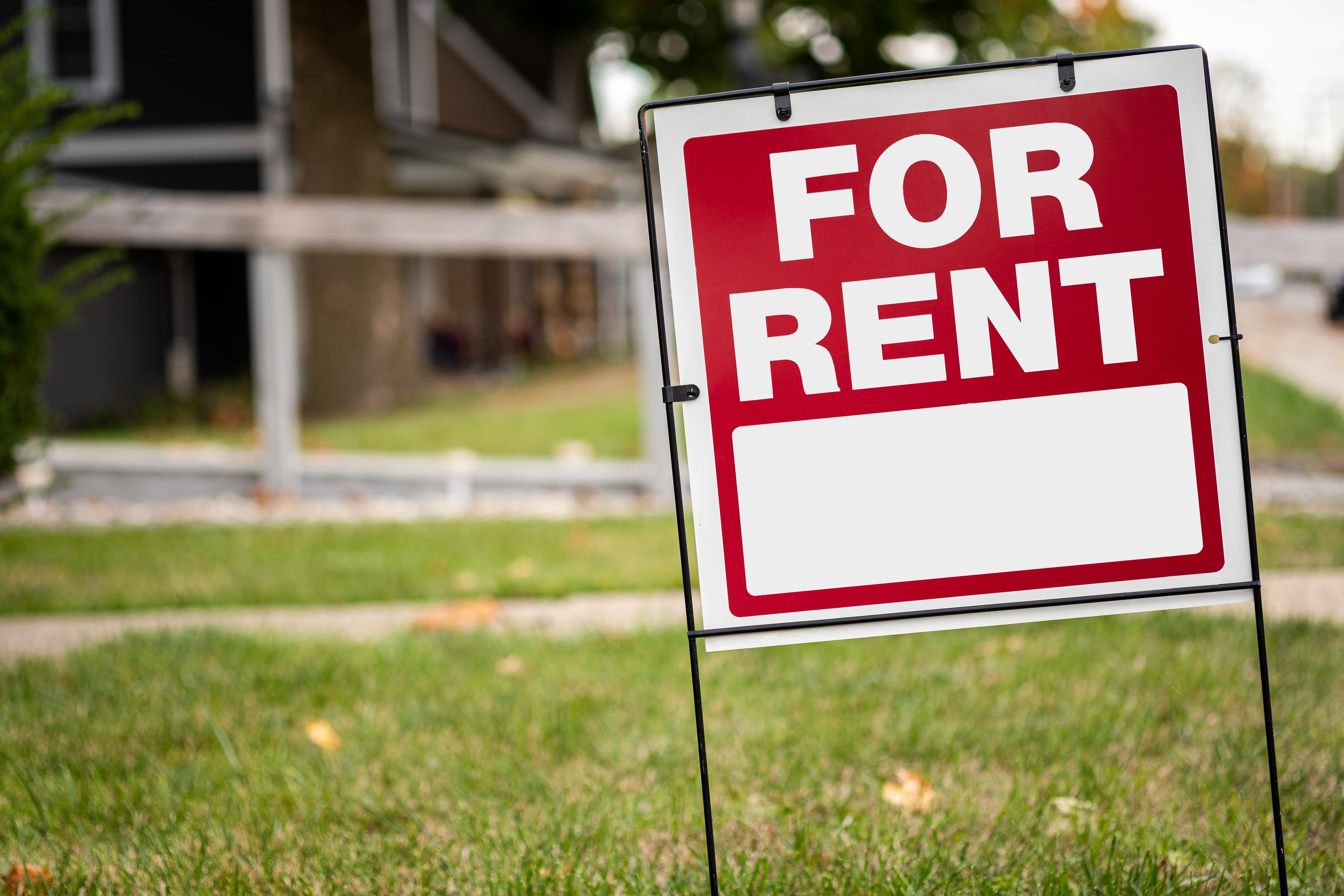 A red and white 'FOR RENT' sign is placed on a grassy lawn in front of a residential house, indicating that the property is available for leasing.