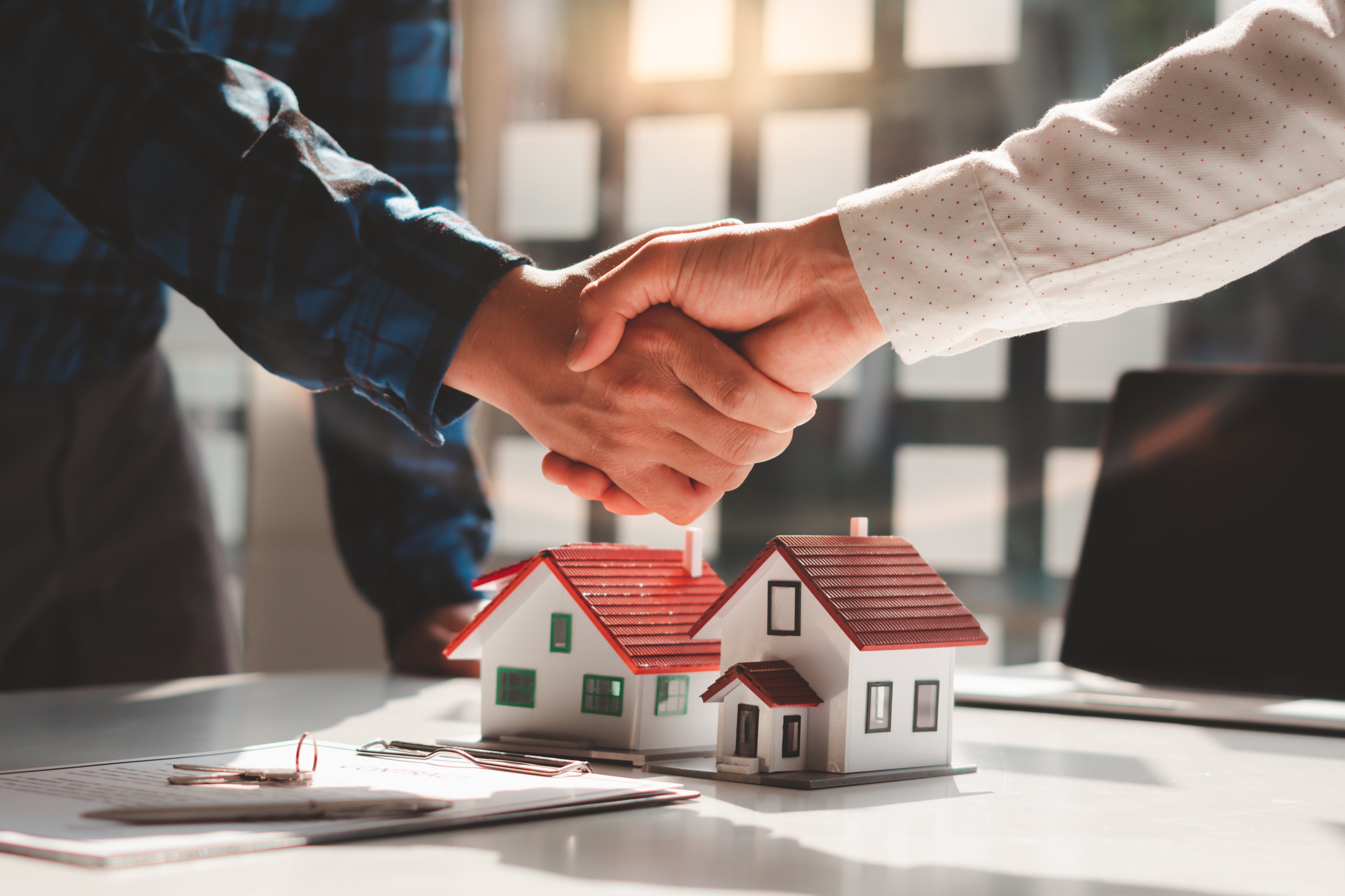 Two people shaking hands over a desk with miniature houses and contract documents, symbolizing a successful real estate deal, property management agreement, or investment partnership.