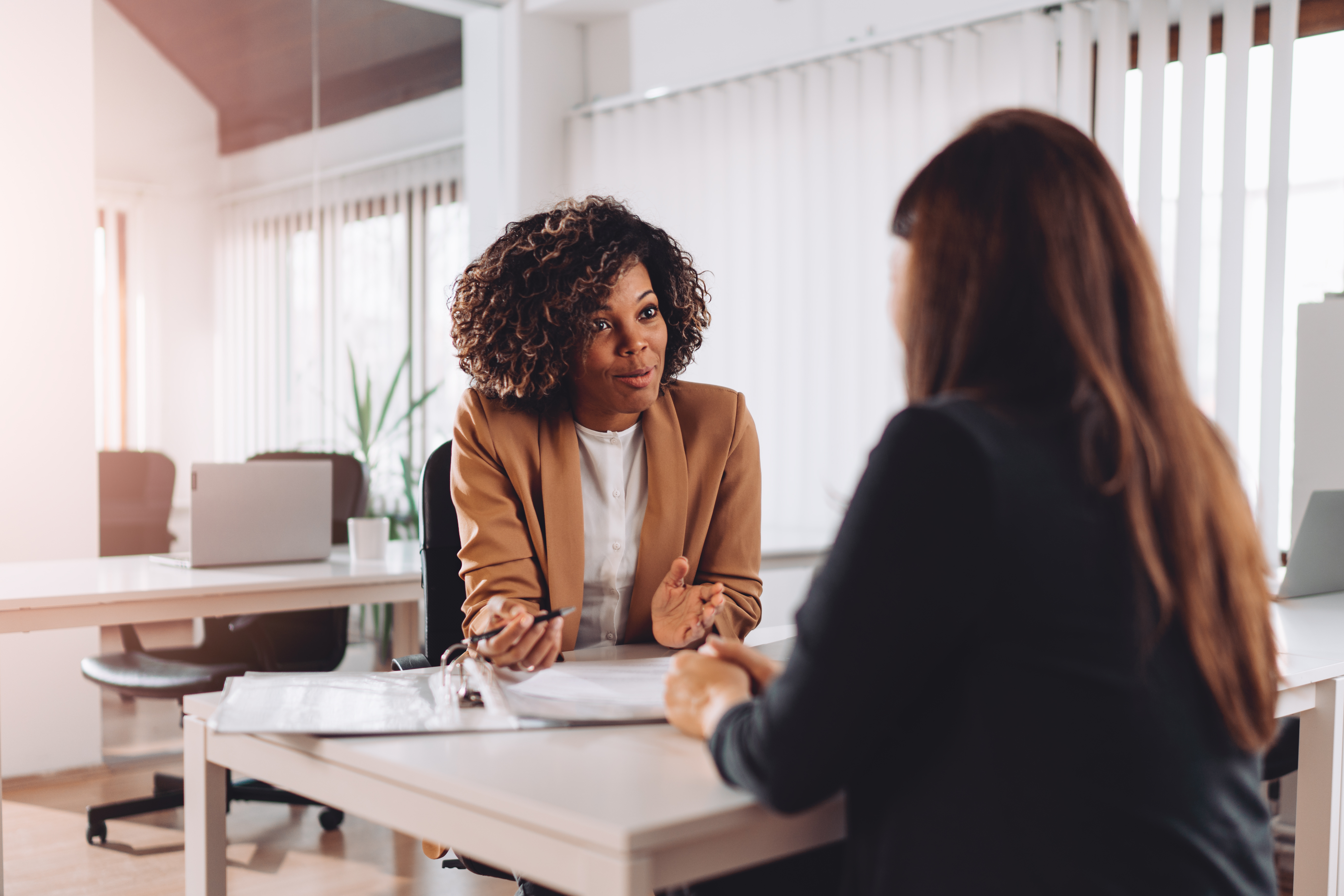 A businesswoman with curly hair and a tan blazer is sitting at a desk, engaging in a conversation with another person in a modern, well-lit office.