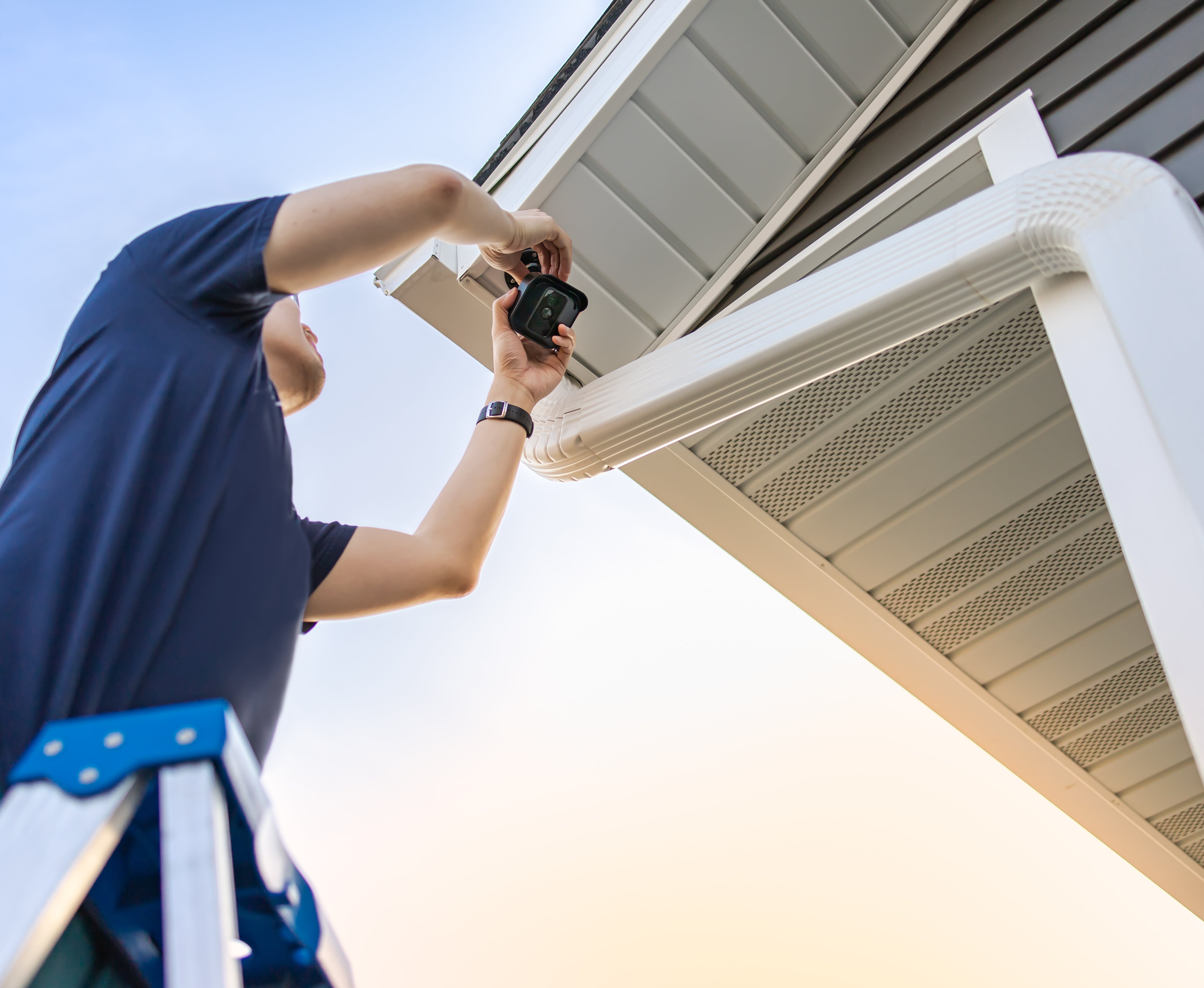 A man on a ladder installing or adjusting a security camera under the eaves of a house. The image represents home improvement, safety, and property maintenance.