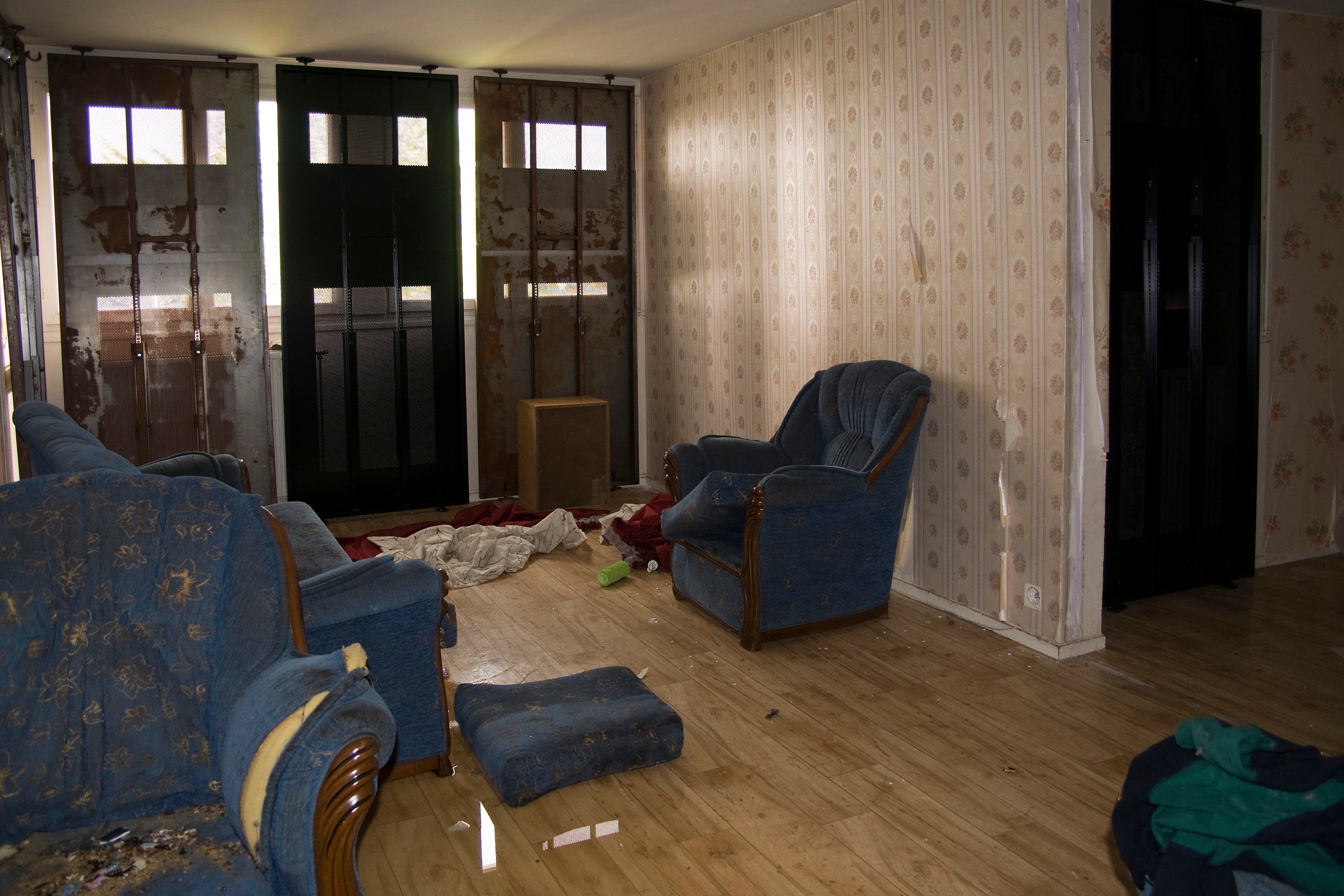 A dimly lit living room with old and dirty furniture. The room shows signs of neglect and vandalism, with peeling wallpaper and boarded-up windows. A blue armchair and sofa are present, both showing signs of wear and tear, along with scattered debris on the floor.