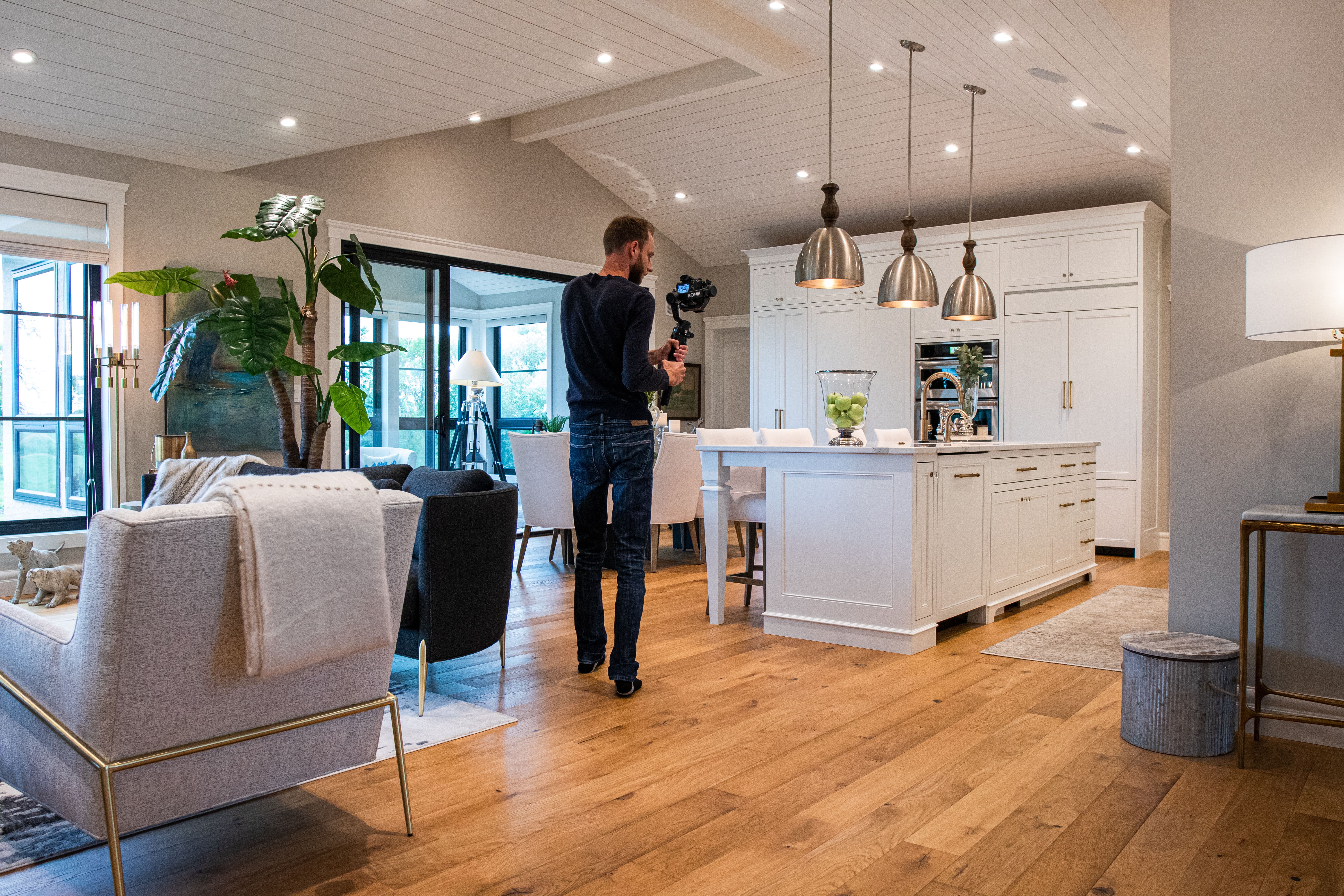 A videographer films the interior of a stylish, modern home with bright lighting, a white kitchen, and wooden floors.