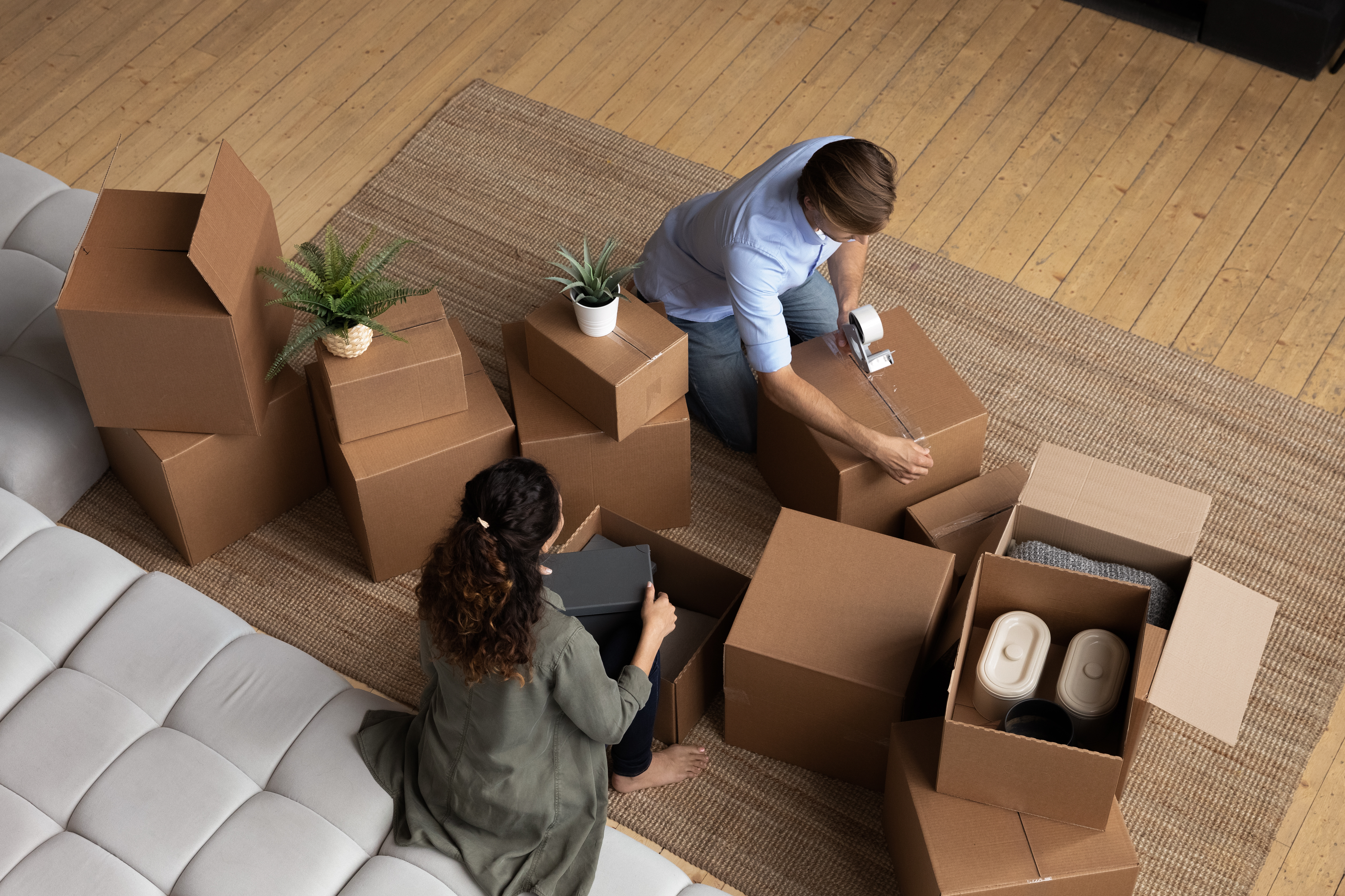  A couple packing cardboard boxes in their living room, preparing for a move. The floor is wooden with a woven rug, and plants are placed on top of some boxes.