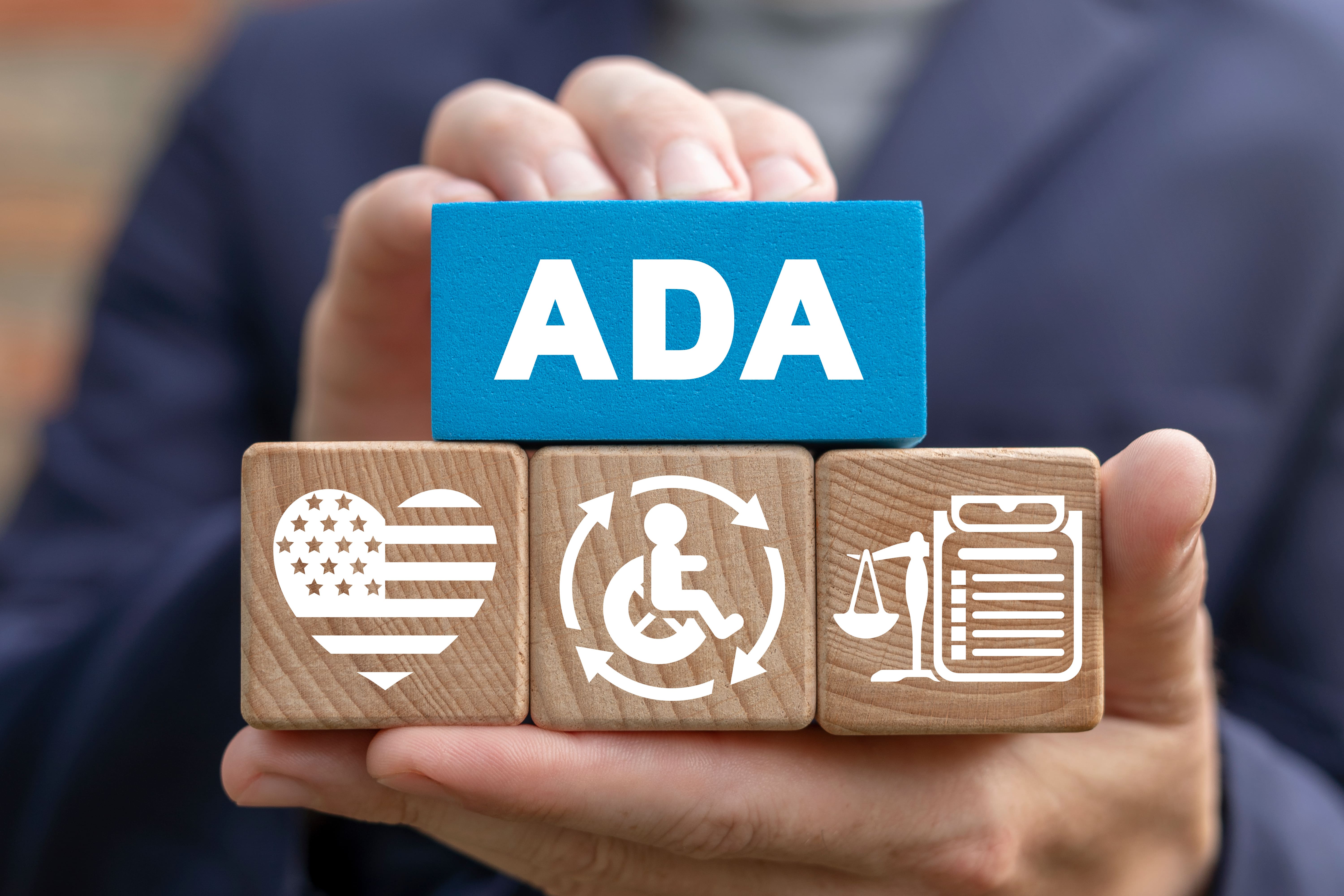 Hand holding wooden blocks with ‘ADA’ and accessibility symbols, representing the Americans with Disabilities Act.