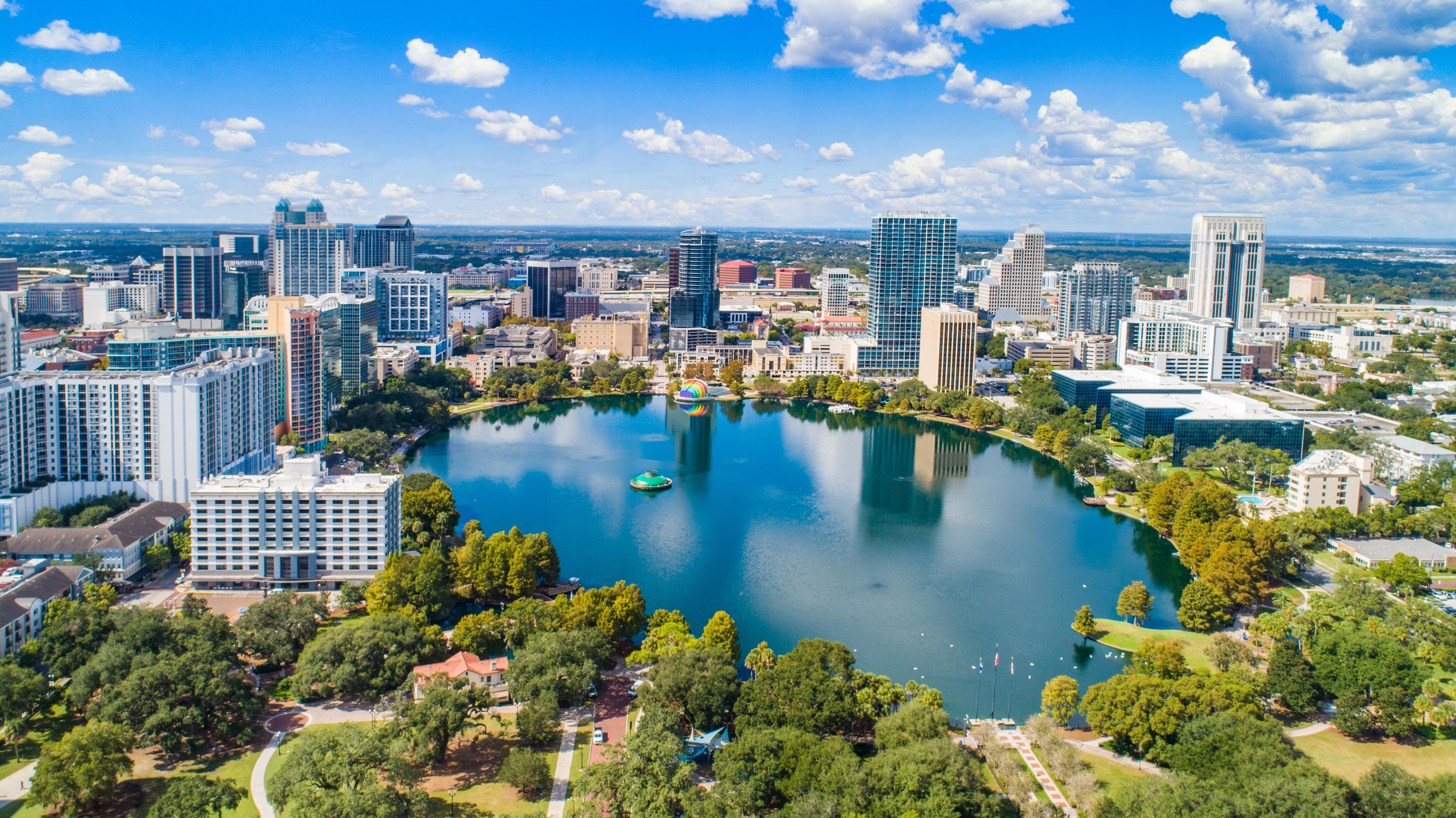 Aerial view of downtown Orlando, Florida, featuring Lake Eola surrounded by high-rise buildings under a bright blue sky.