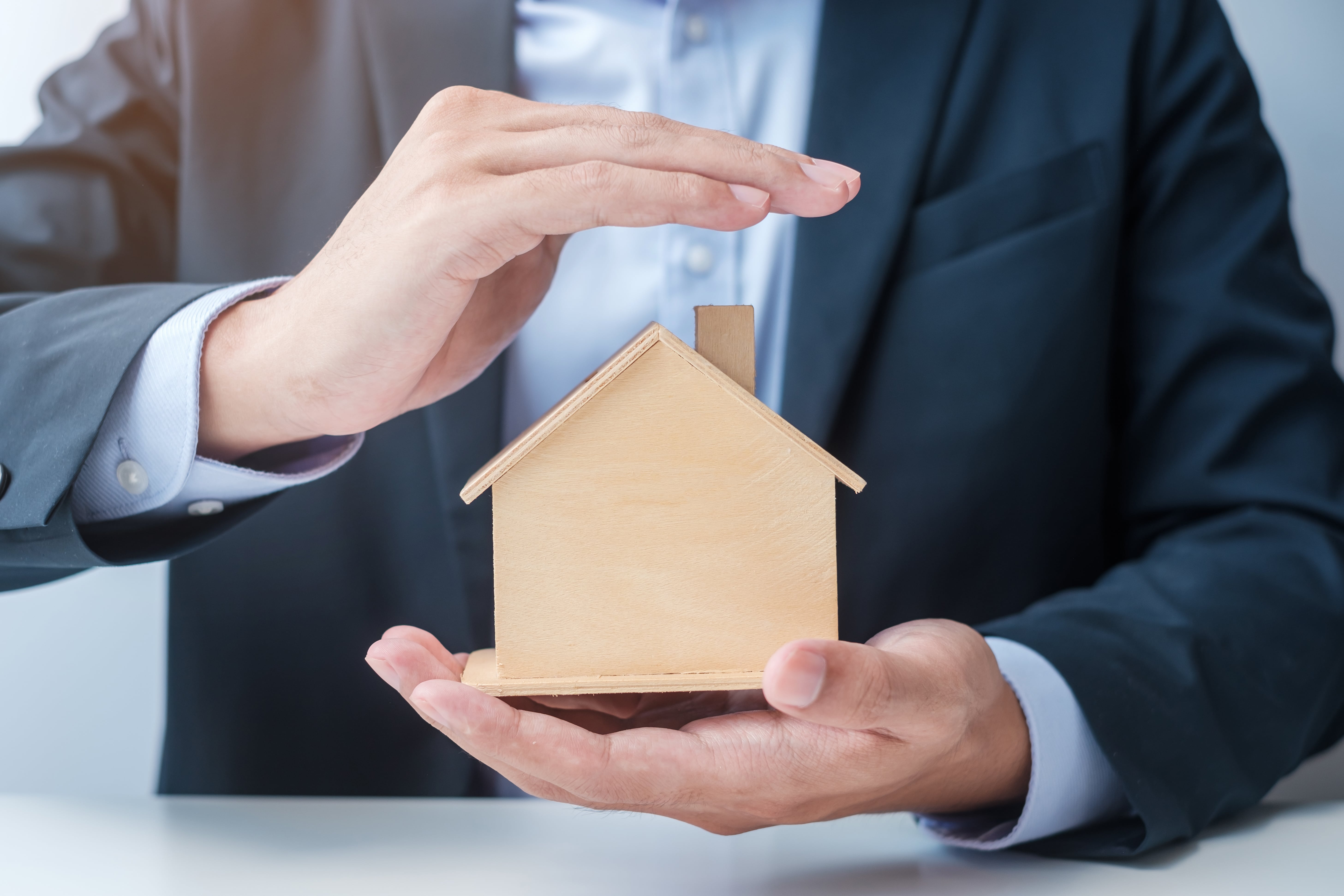 A person in a business suit gently covering a small wooden house model with their hands, representing protection, property management, and real estate investment security.