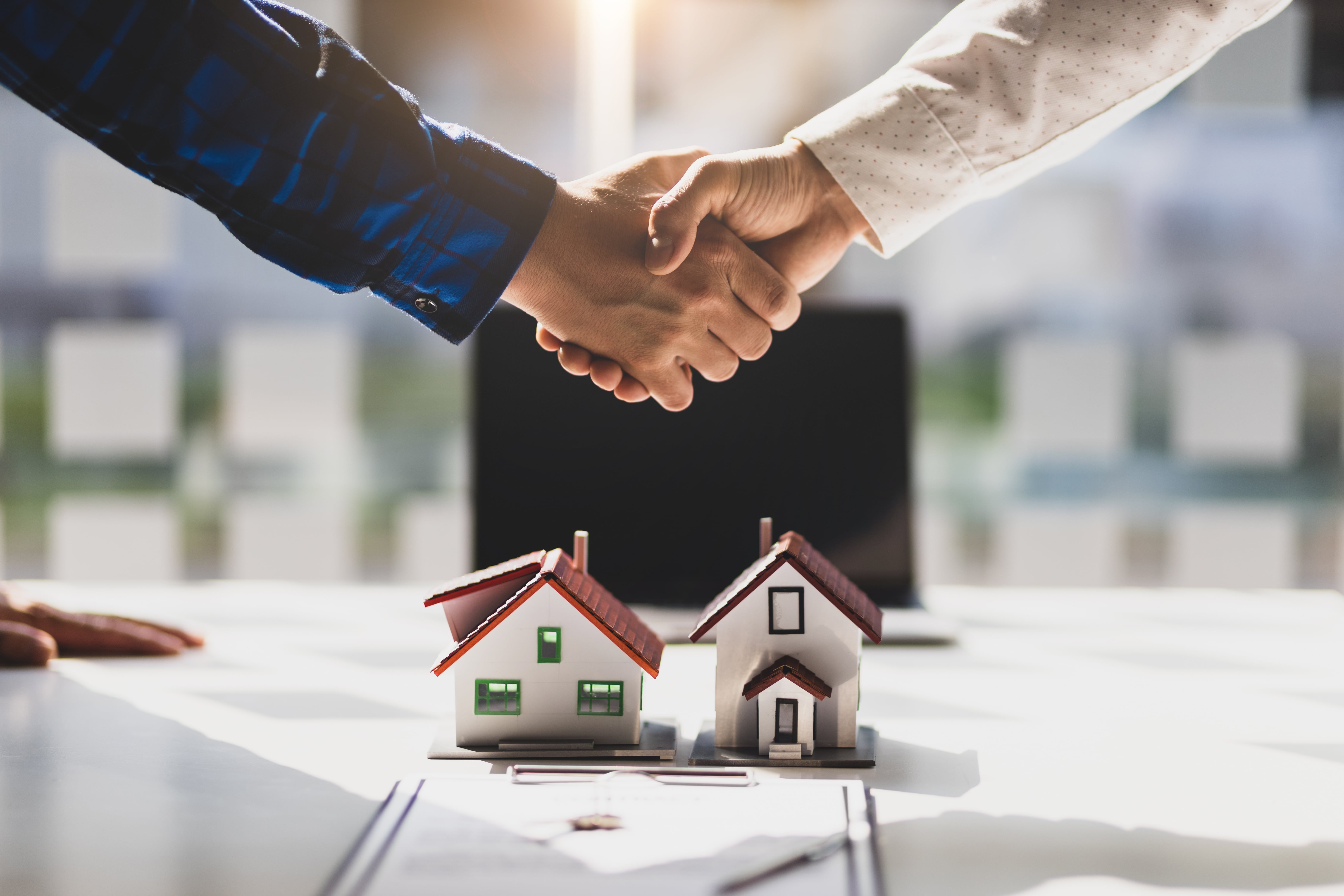 Two people shaking hands over a desk with small model houses and documents, symbolizing a successful real estate transaction, property investment, or business agreement.