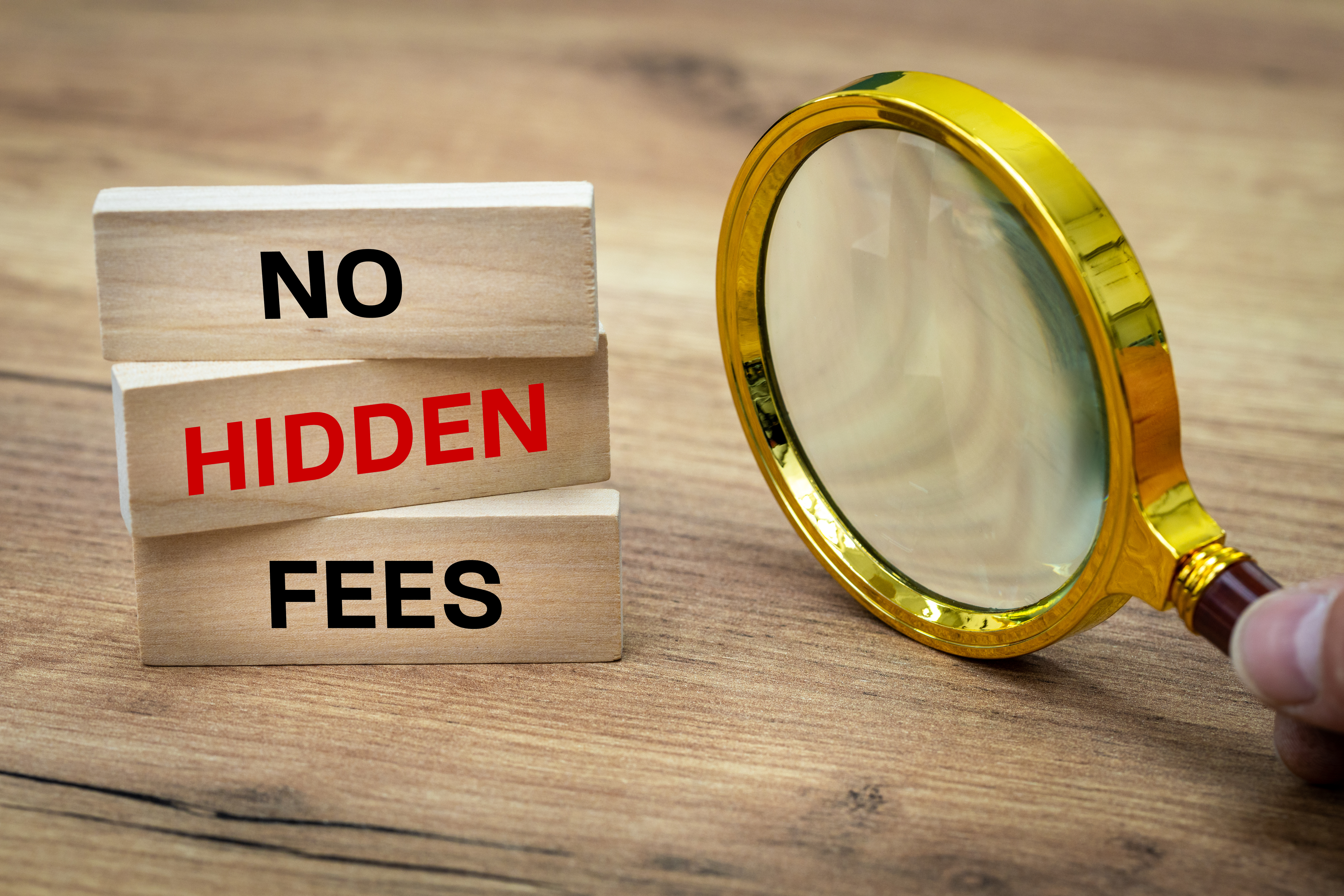 Wooden blocks stacked vertically with the words “No Hidden Fees” in black and red text. A gold magnifying glass is positioned next to the blocks, symbolizing scrutiny and transparency. The scene is set on a wooden surface.