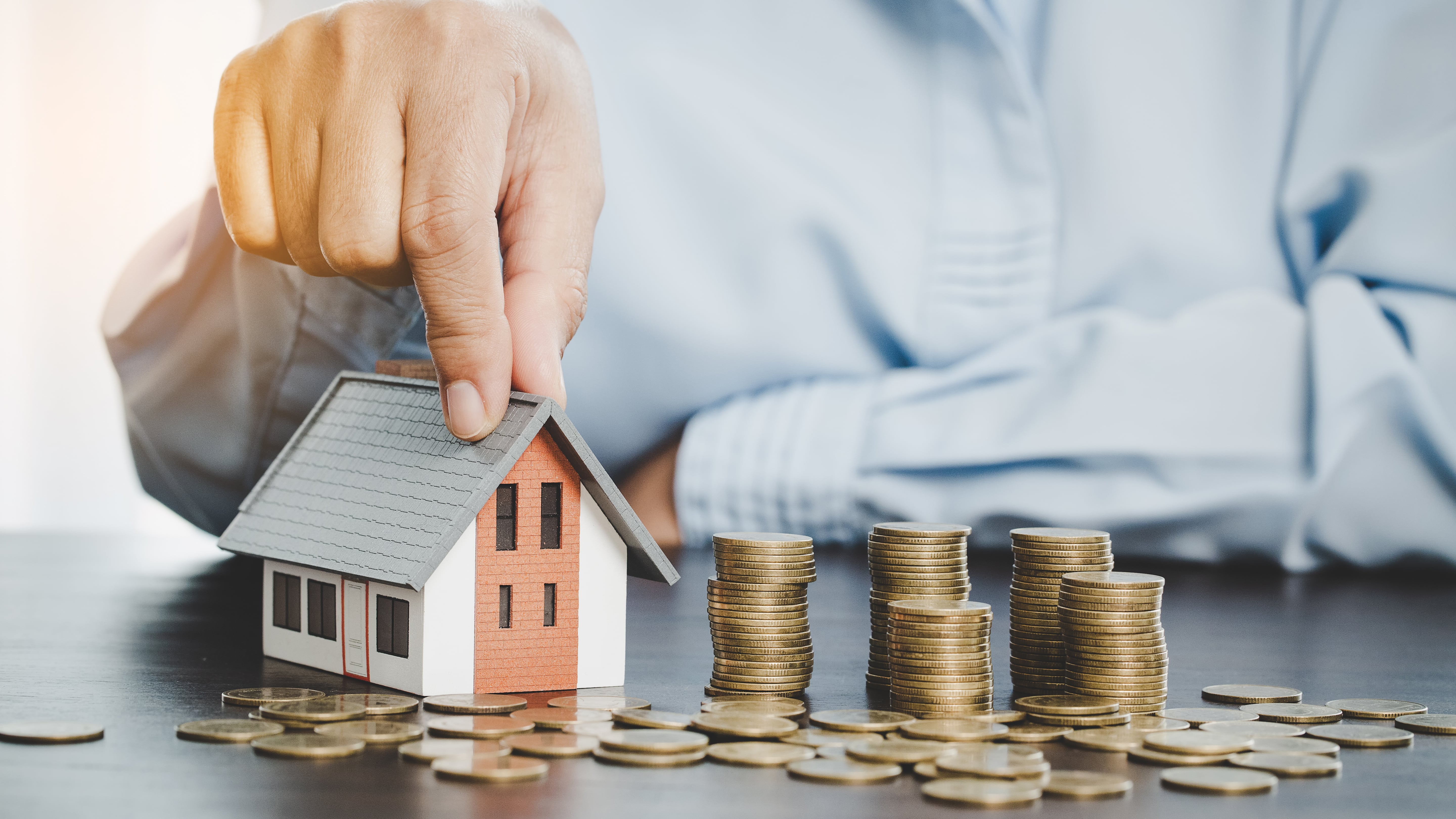 A hand placing a model house next to stacks of coins, representing property investment and rental income growth.