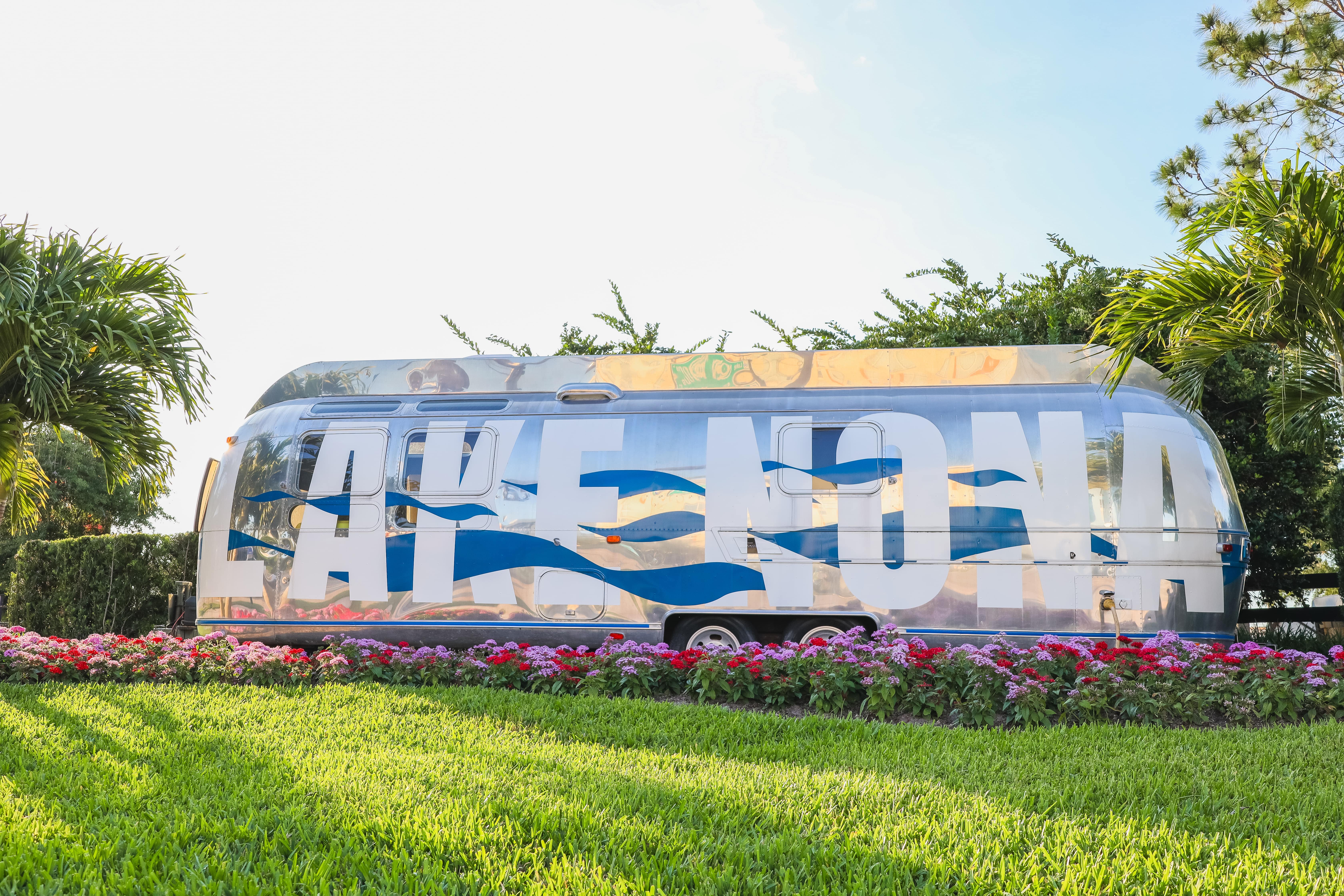 A sleek, silver Airstream trailer with 'Lake Nona' written in bold blue and white letters, surrounded by lush greenery and vibrant flowers. The reflective surface of the trailer captures the bright sky and palm trees.