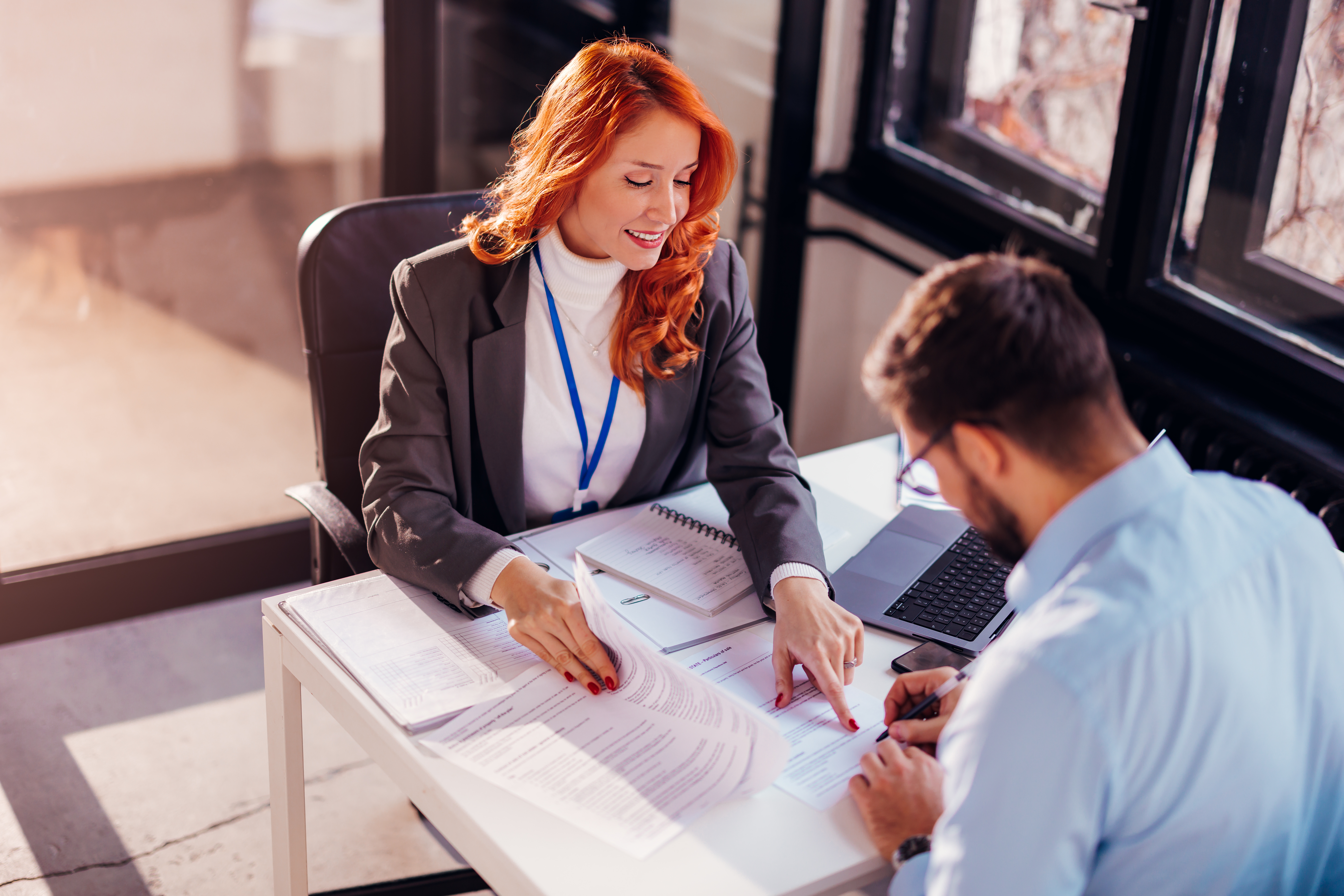 A professional woman with long red hair, wearing a gray blazer and white turtleneck, is smiling while discussing paperwork with a client across the table. The office environment is bright, with natural light pouring in from large windows.