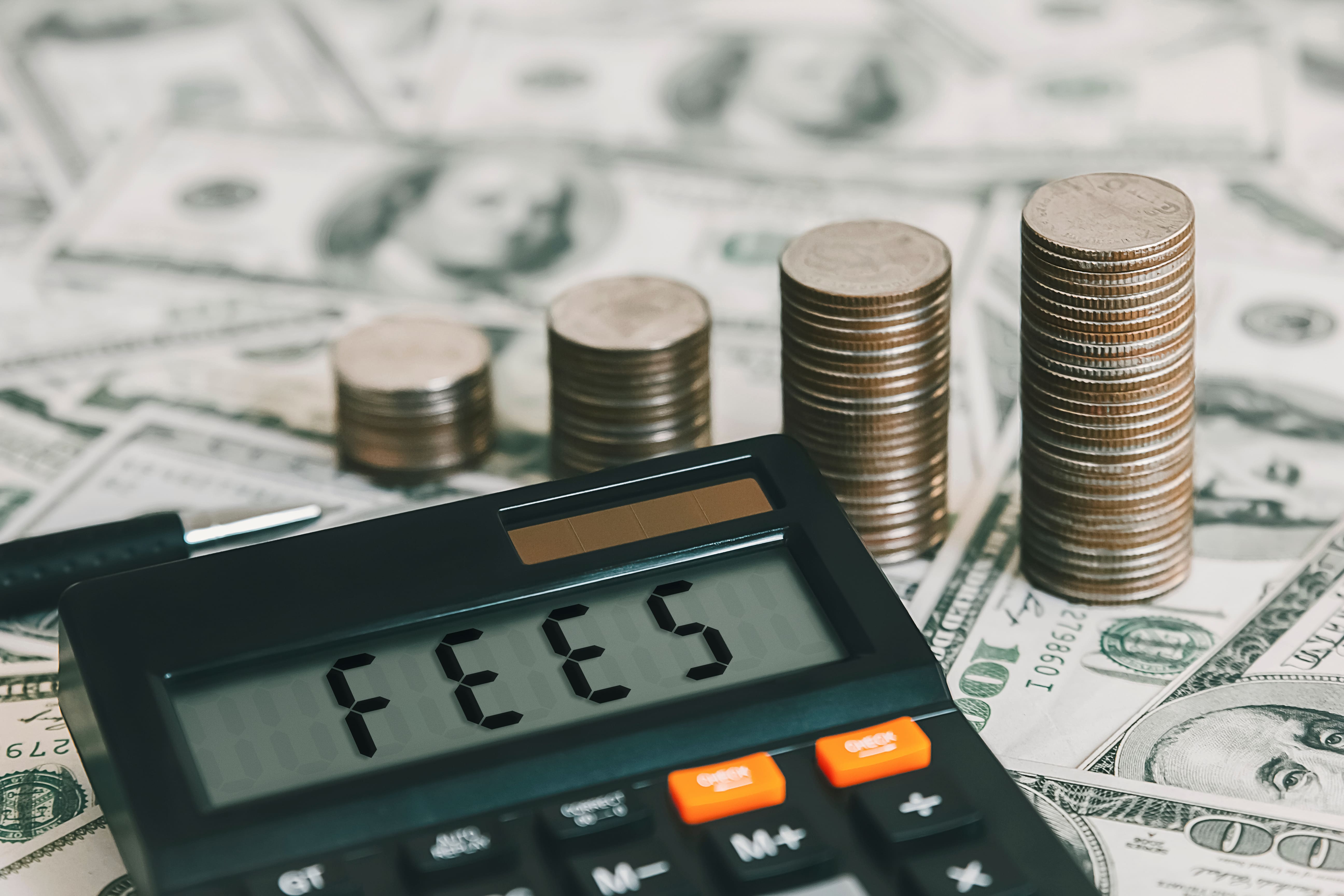 A calculator displaying the word 'FEES' sits on a table covered with dollar bills, with stacks of coins increasing in height in the background, symbolizing financial management and costs.