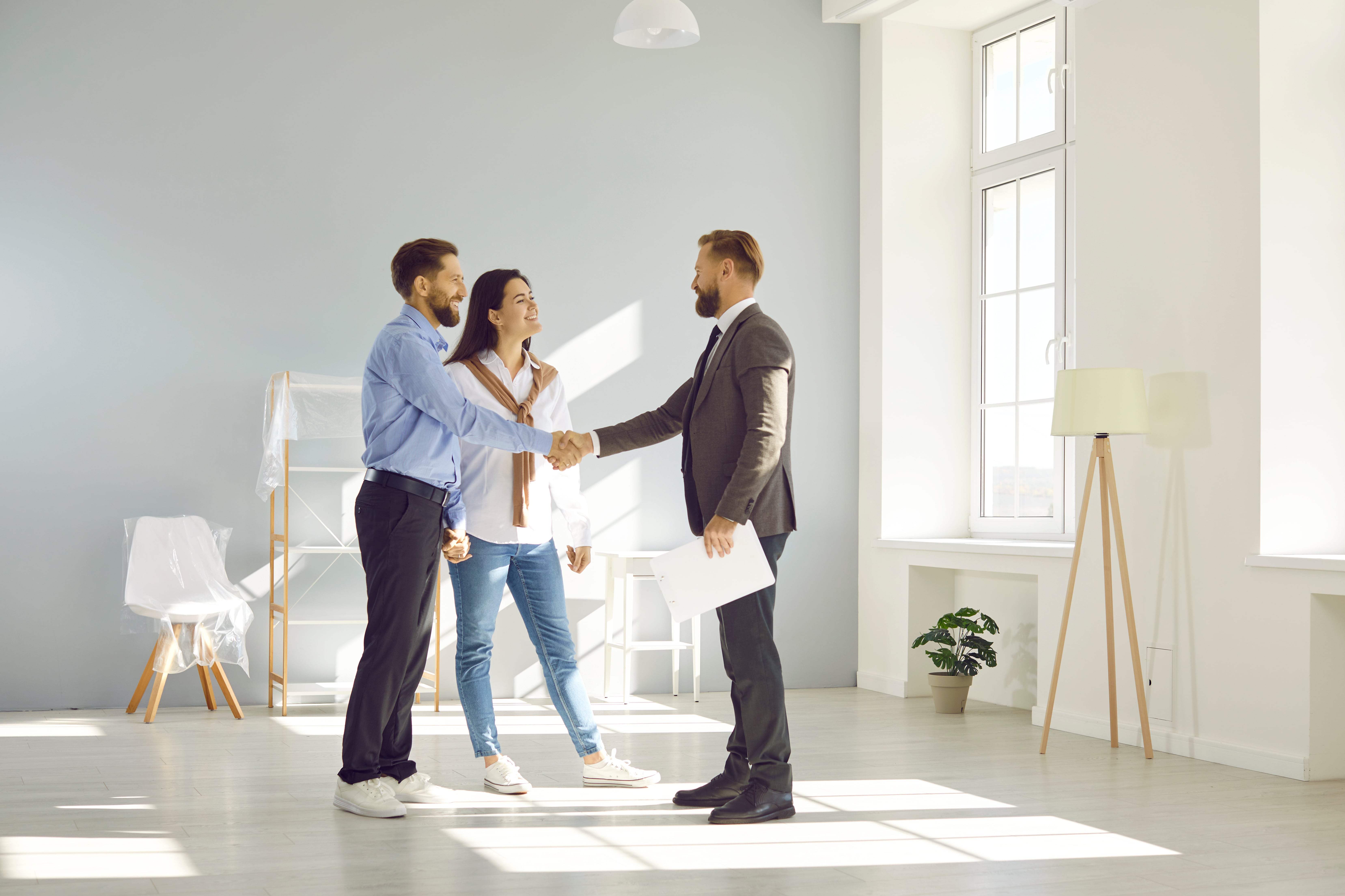 A professional real estate agent shaking hands with a couple in a bright, modern home. The couple looks happy, indicating a successful home-buying or rental process.