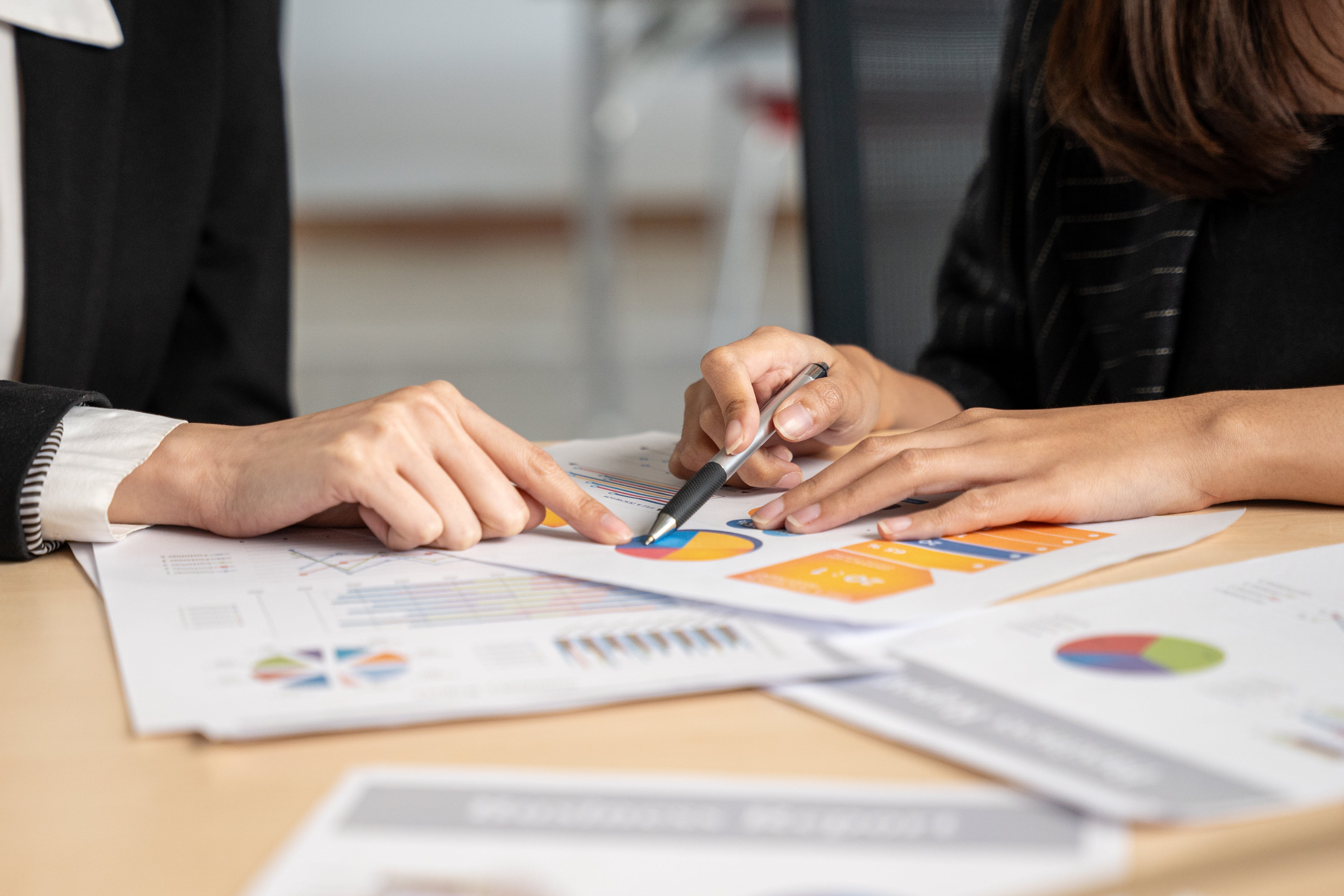 Two professionals in business attire review colorful charts and graphs on printed documents during a financial or business meeting.