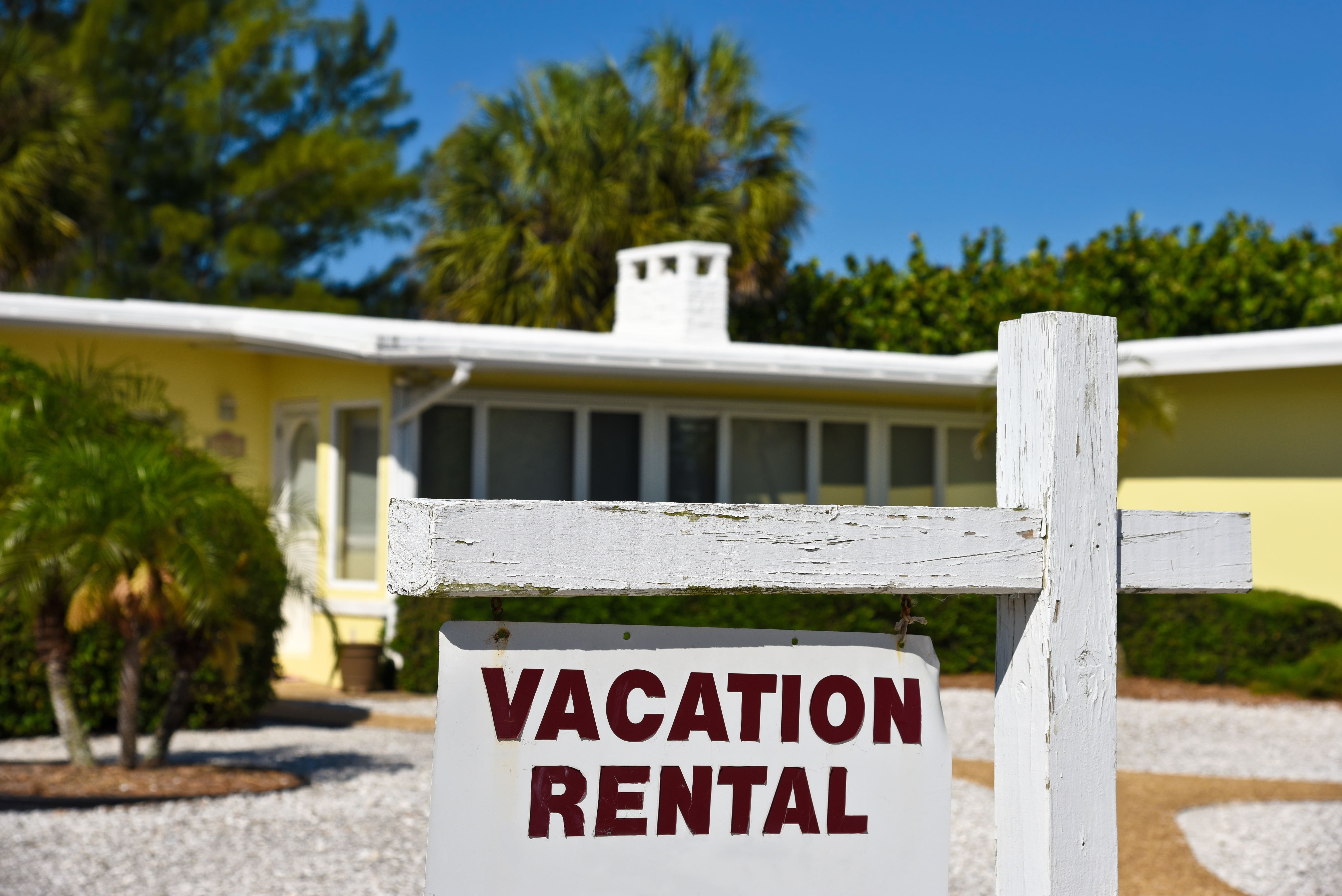 Close-up of a sign that reads ‘Vacation Rental’ in front of a yellow house with palm trees in the background.