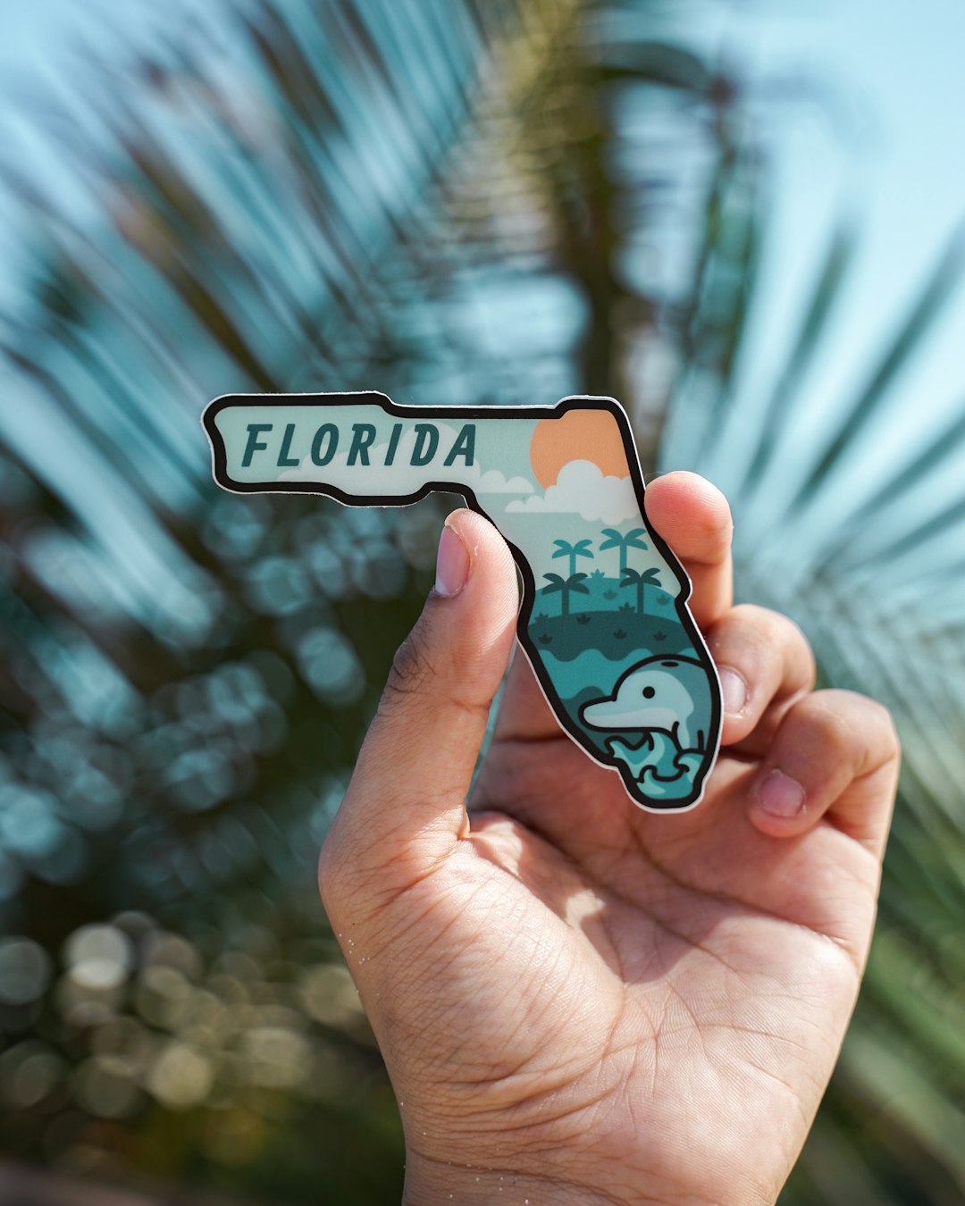 A hand holding a cutout of the state of Florida with a colorful design featuring palm trees, a sun, and a dolphin, against a blurred background of palm leaves and a blue sky.