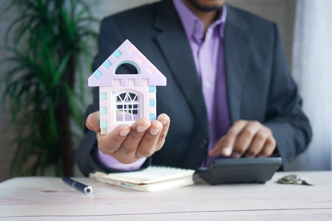 A businessman in a suit holding a small, colorful model house in his hand while using a calculator with the other hand. The background shows a notepad, pen, and some keys on the table, suggesting a real estate or financial setting.