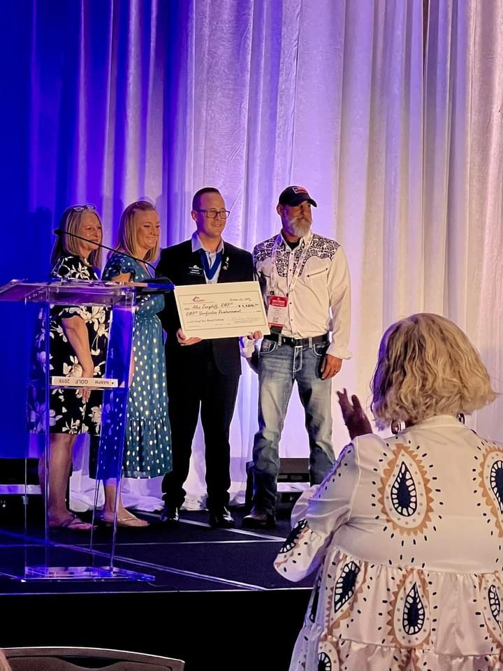 Alex Zweydoff receiving an award on stage with three other people, with an applauding audience in the foreground.