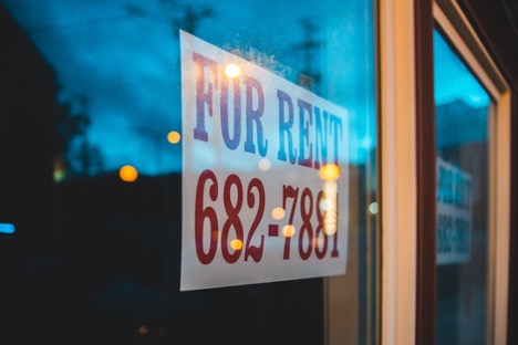 A close-up of a 'FOR RENT' sign displayed on a window with a blurred evening scene in the background.