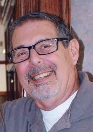 Smiling man with glasses and a gray shirt standing indoors, looking at the camera. A photo of Darrel Kazen.