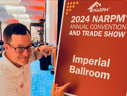 Phil Owen of Onsight Pros pointing to a sign that reads ‘2024 NARPM Annual Convention and Trade Show, Imperial Ballroom,’ in a convention setting.